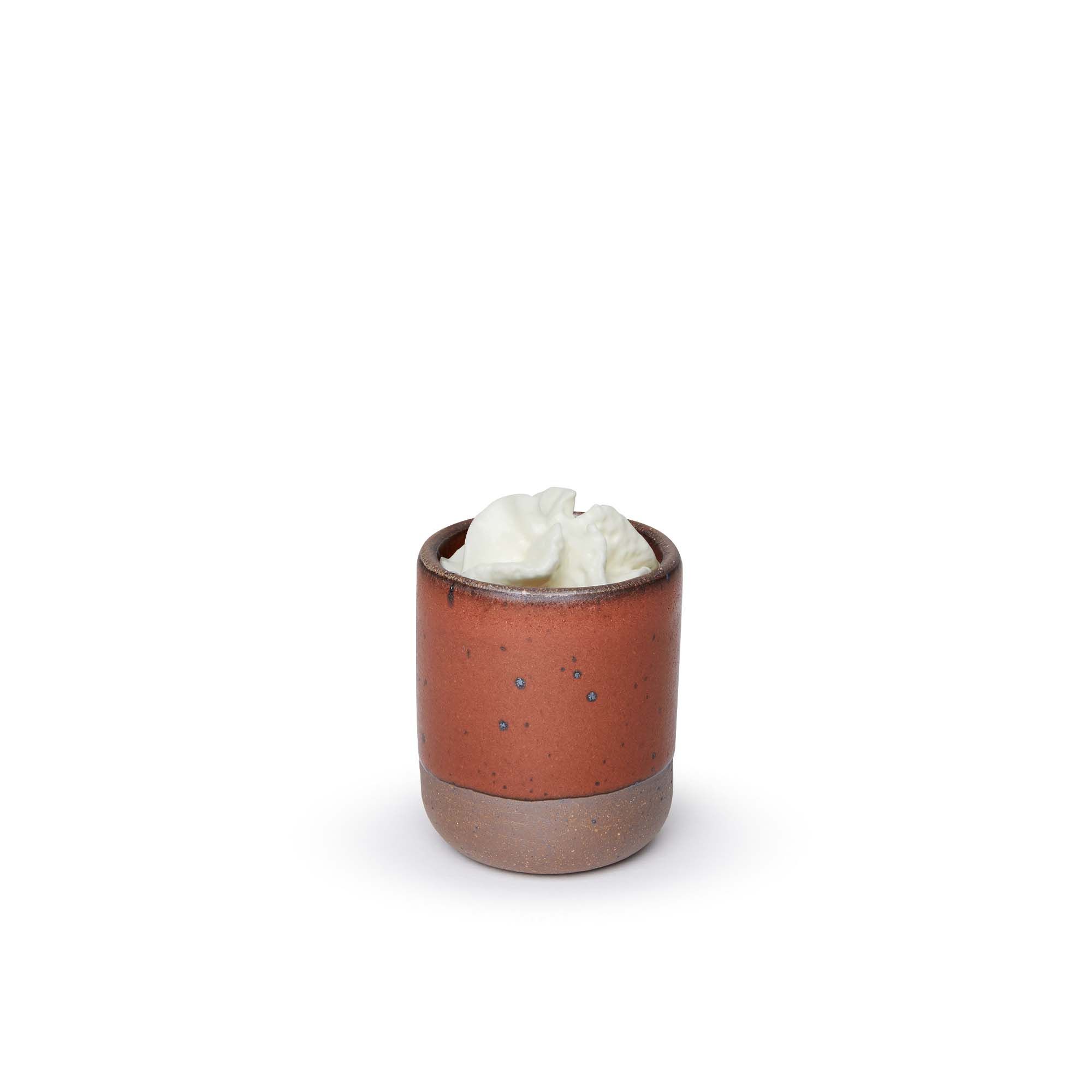 A small, short ceramic mug cup in a cool burnt terracotta color featuring iron speckles and unglazed rim and bottom base, topped with whipped cream.