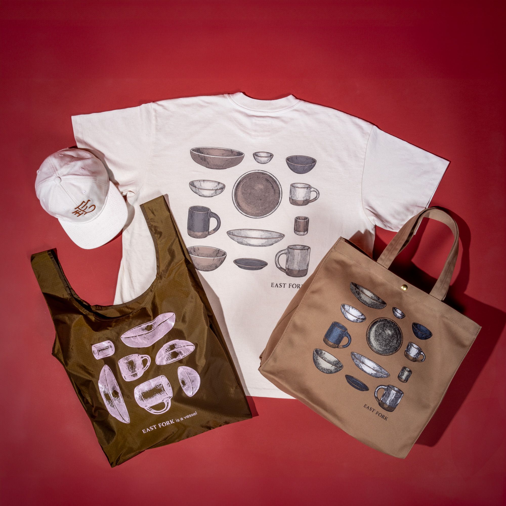 Overhead view of an East Fork tee with ceramic mugs, plates, and mug illustration, with a tote bag, a nylon bag, and a baseball hat - all against a red background.