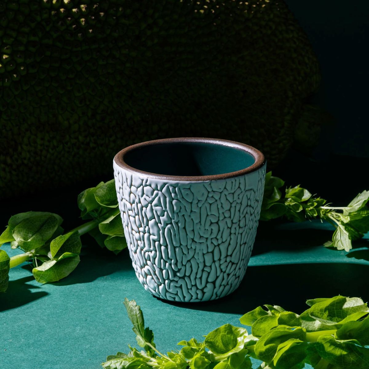 A small ceramic cup with cracked texture and the interior being dark teal, sitting in a moody studio setting with foliage