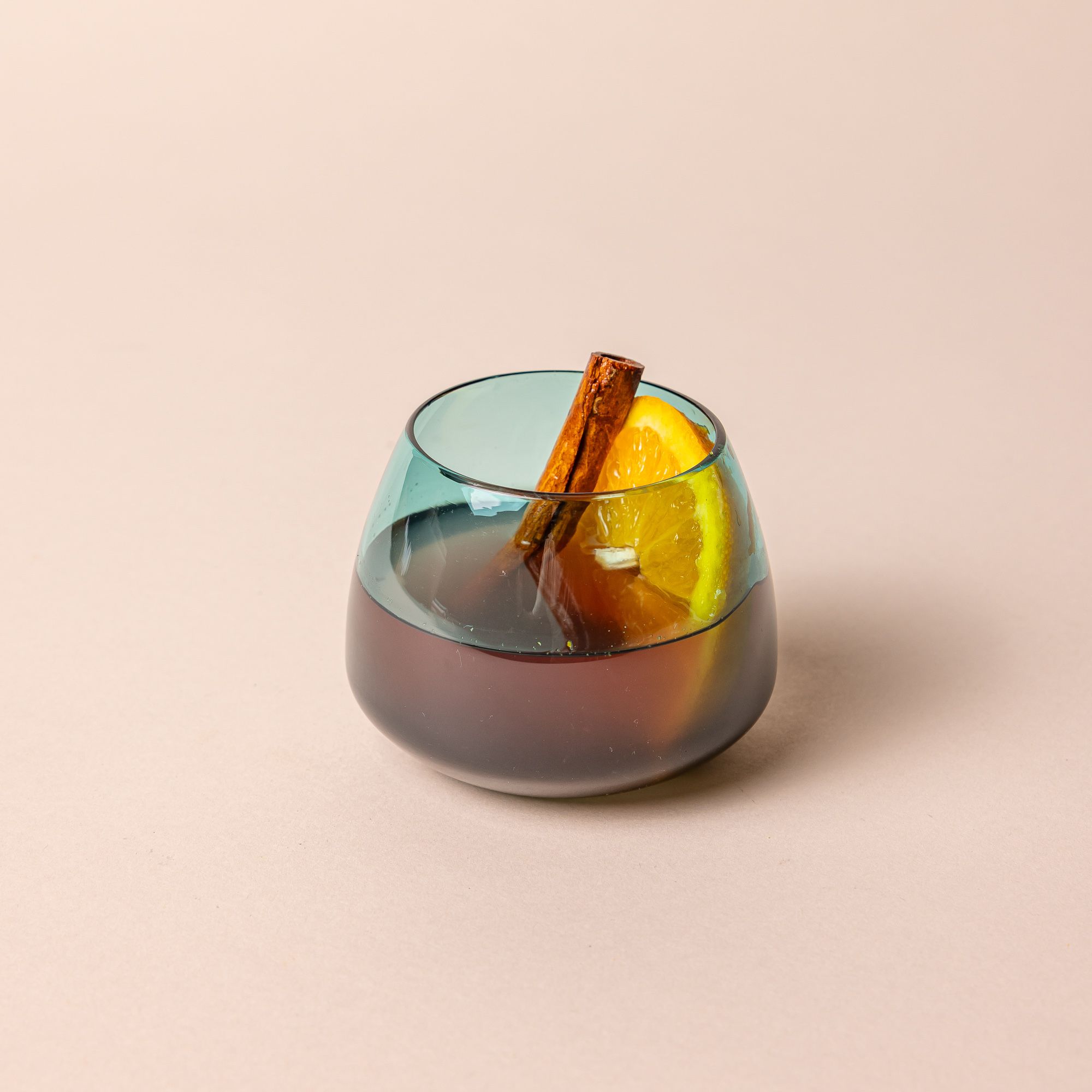 A round short aqua blue glass punch cup filled with a winter cocktail featuring an orange slice and cinnamon stick. The cup has a round base that narrows a bit near the top.