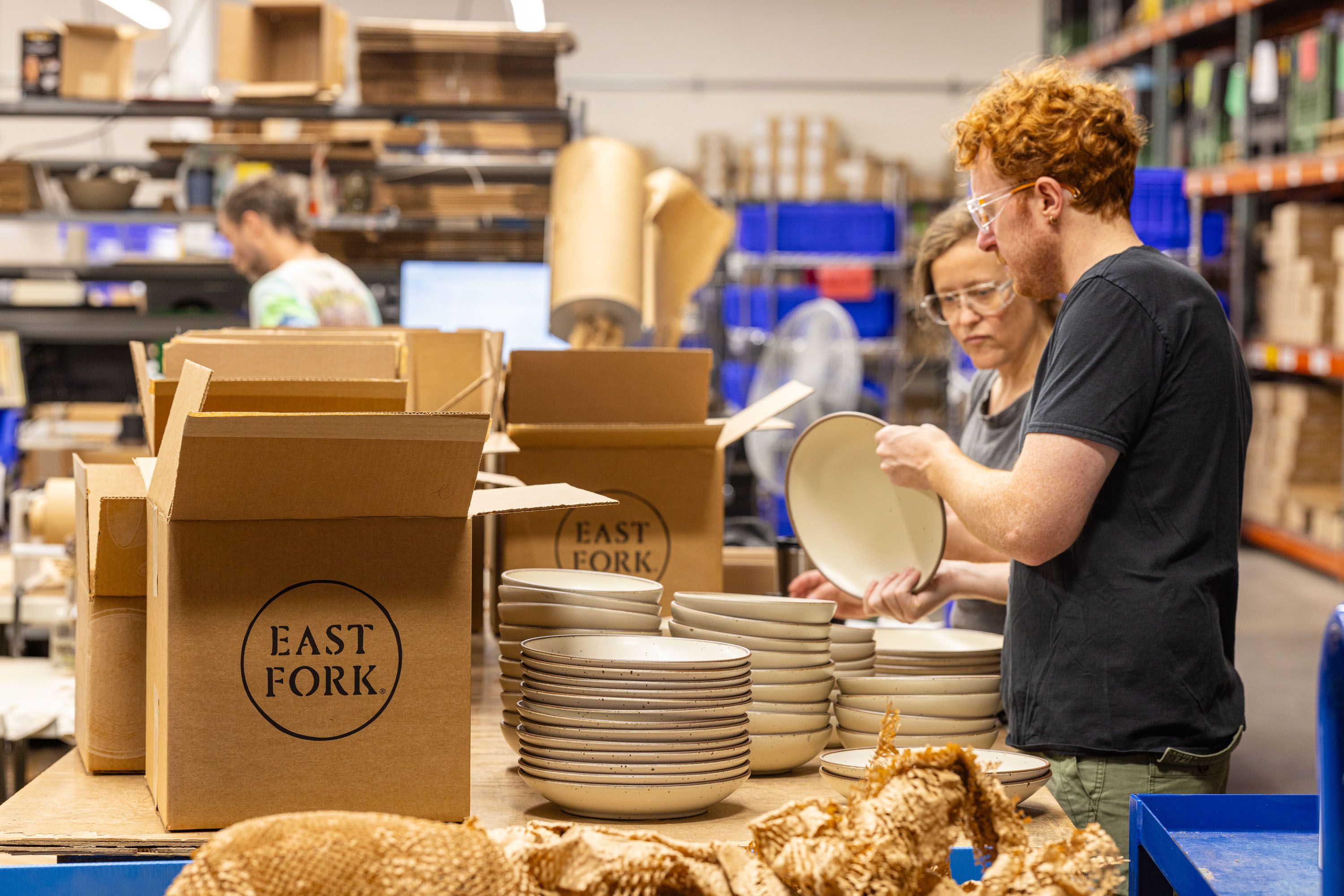 In a factory setting, people are inspecting ceramic plates and shallow bowls in a cream color, surrounded by packaging material and a box that reads "East Fork"