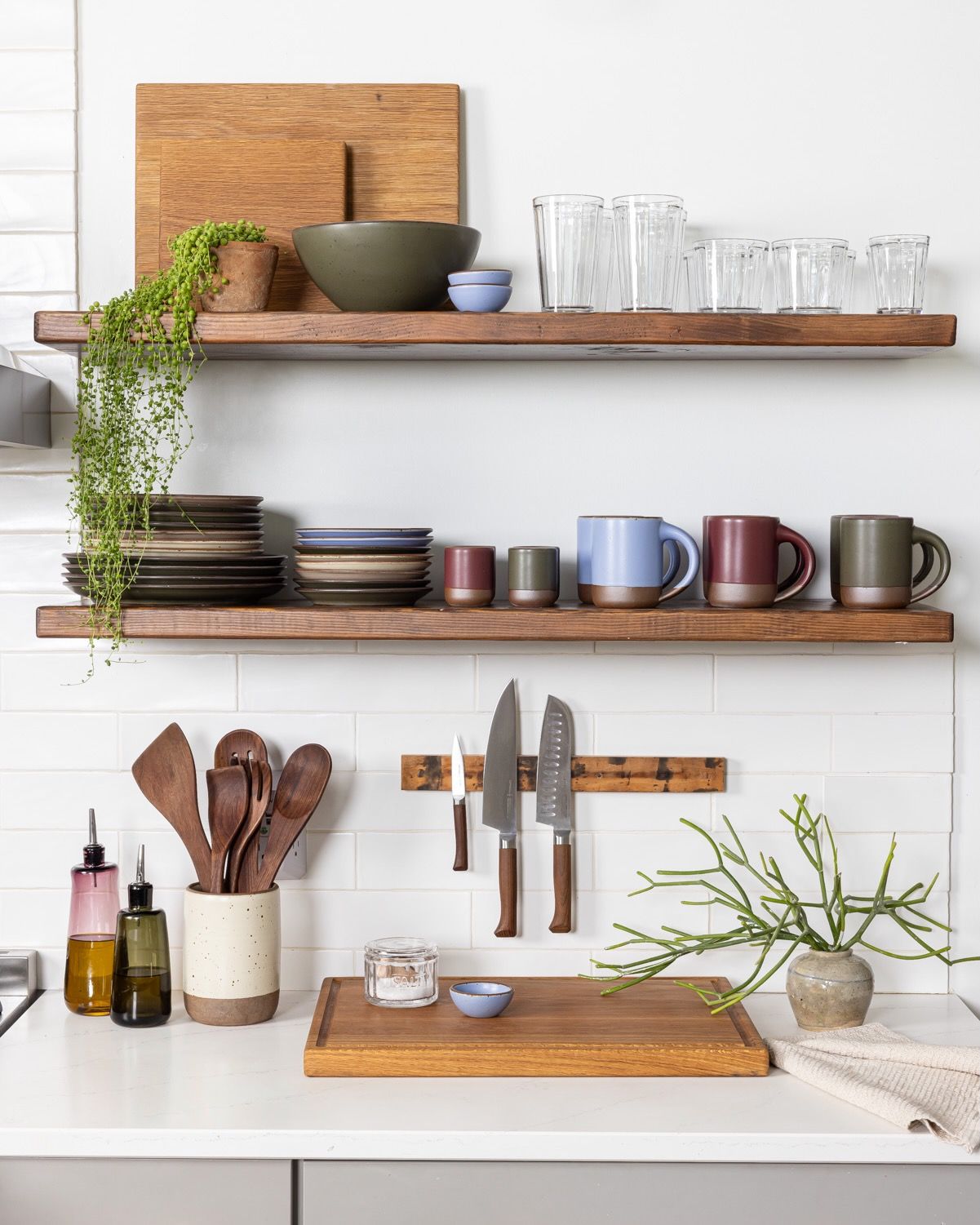 A kitchen shelf filled with ceramic plates, cups and mugs in forest green, periwinkle, and plum along with simple diner glasses, a cutting boards, and other kitchen accessories.