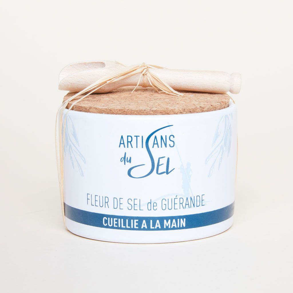A wooden scoop on top of a white and blue container for fleur de sel that has a cork top 