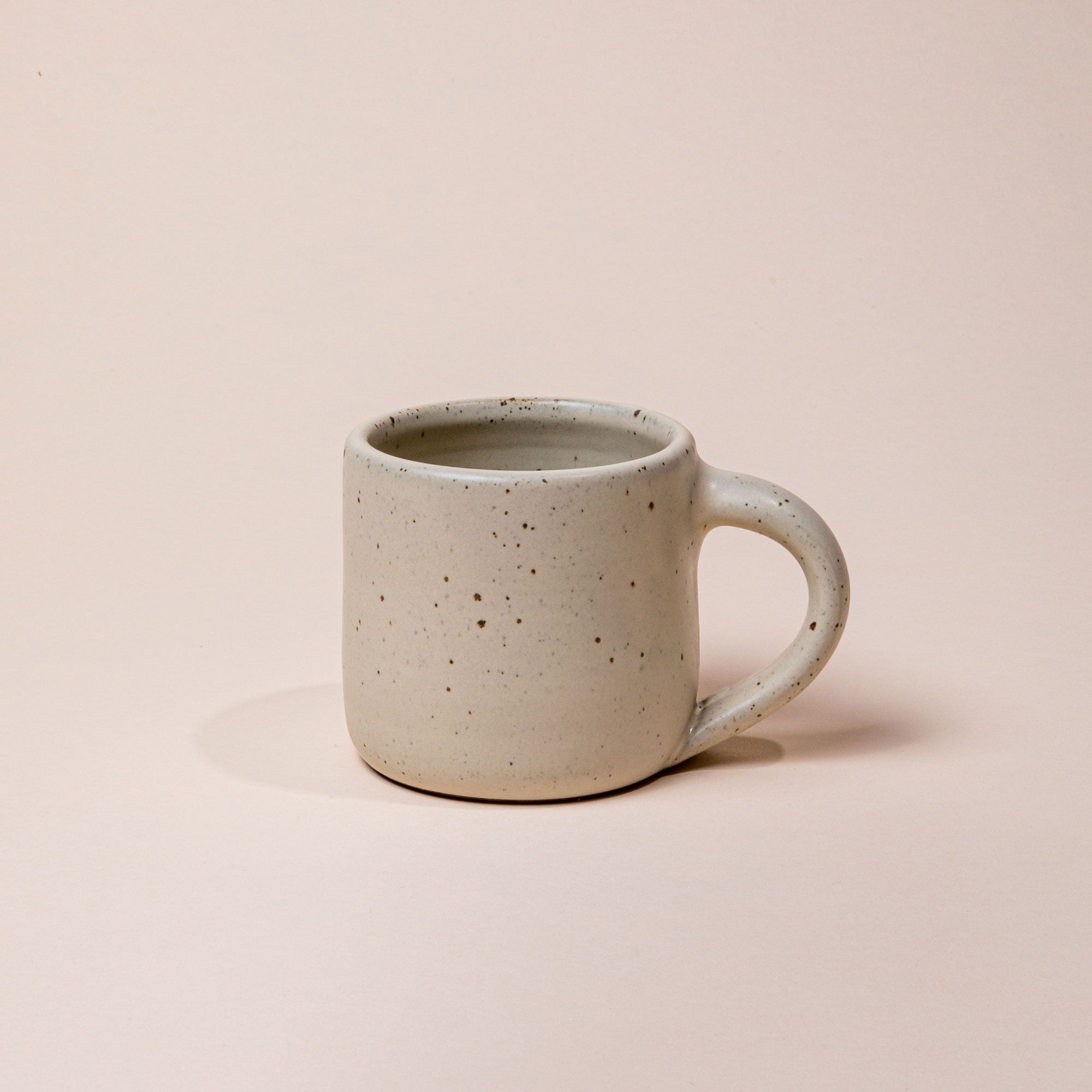A medium sized ceramic mug with handle in a warm, tan-toned, off-white color featuring iron speckles