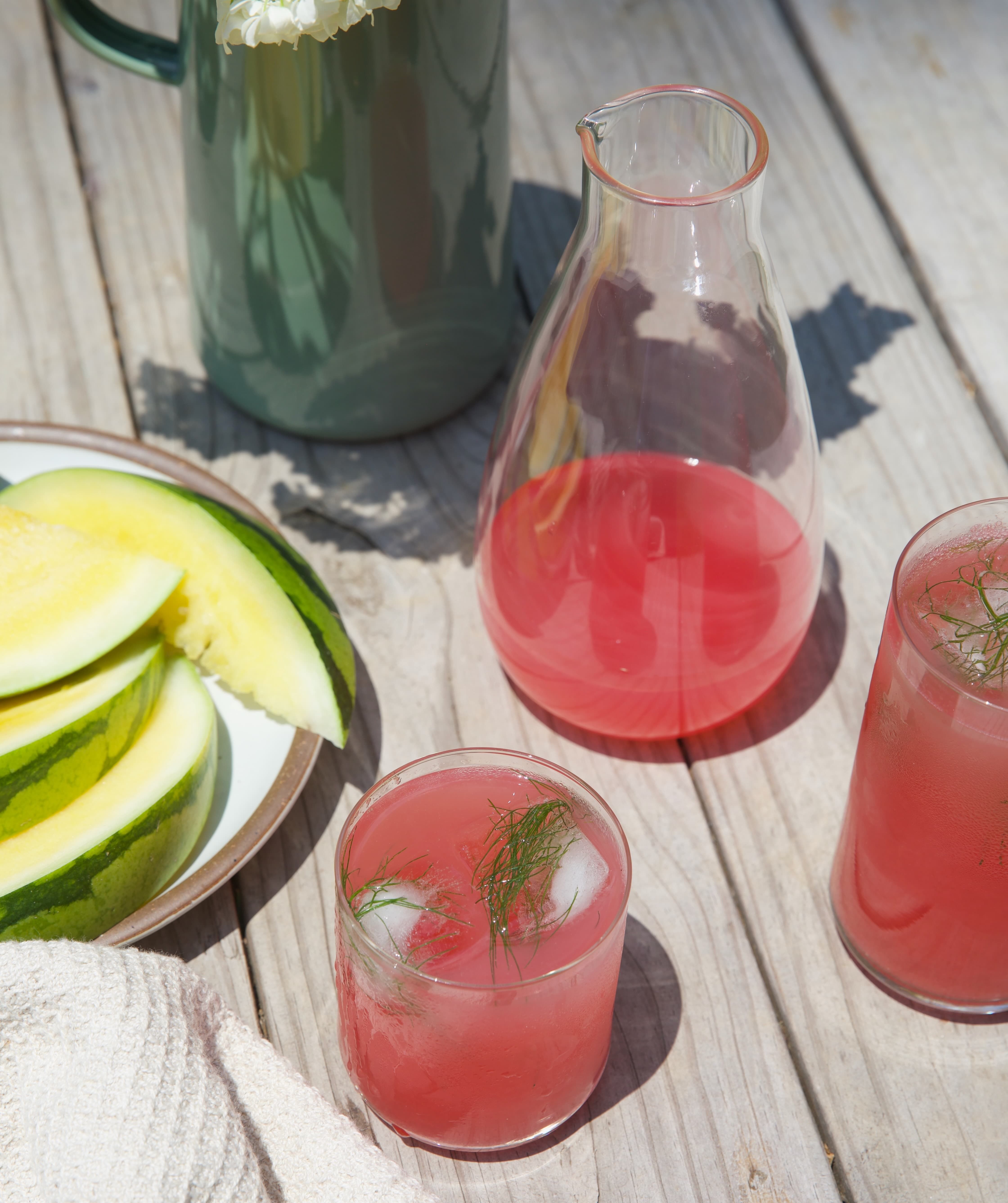 On a picnic table is 2 glasses and a carafe filled with a pink drink with ice. There is also a plate of sliced watermelon.