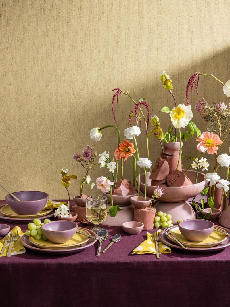 A beautiful tablescape filled with zesty yellow and soft mauve purple ceramic dinnerware, purple tablecloth, and large and lively floral sculpture centerpiece
