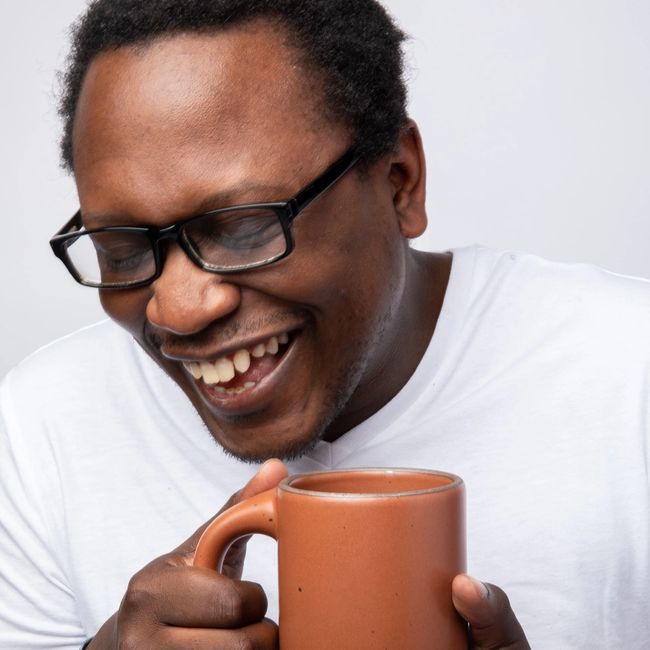A person wearing glasses is smiling and looking down while holding an East Fork mug close to their face in Amaro, a cool terracotta color.