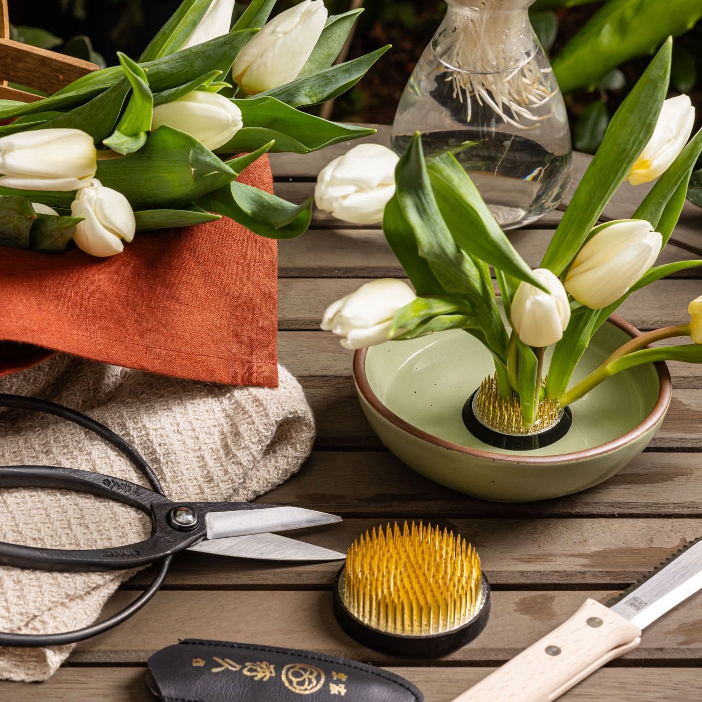 On a table are gardening and floral accessories, including a flower frog by itself, a flower frog with tulips on it in a shallow bowl with water, garden snips, a gardening knife, and a basket of tulips.