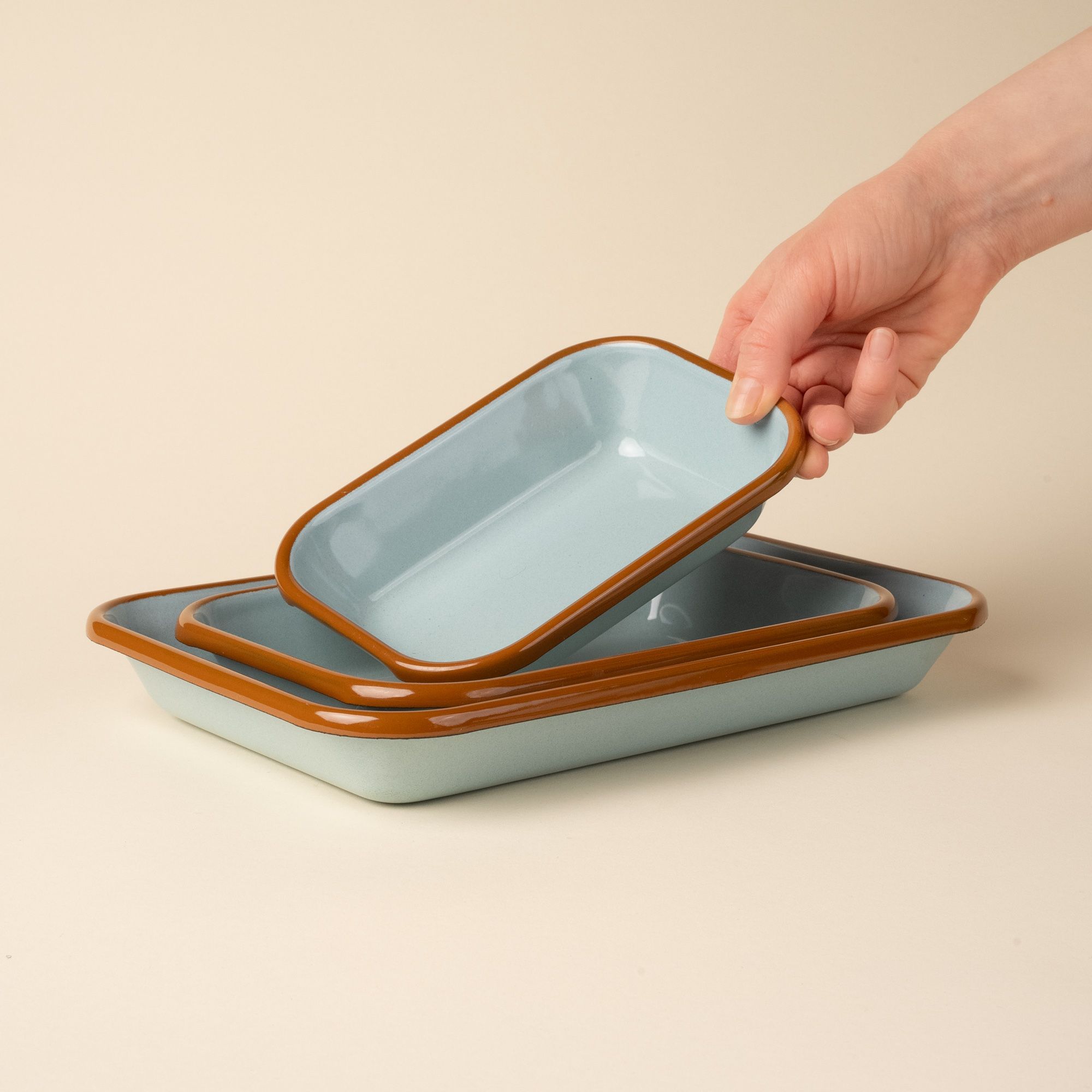 A hand placing a small dish into a nesting trio rectangular baking dishes in a robin's egg blue color with a warm brown rim.