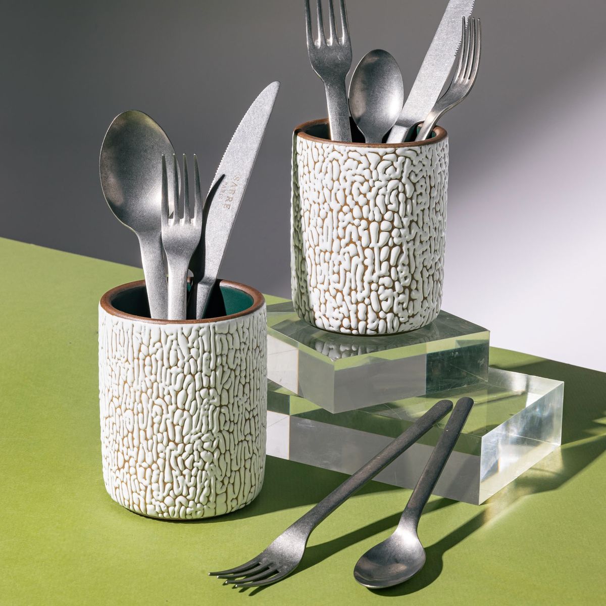 Two medium white ceramic vessel with cracked texture and the interior being dark teal, filled with steel flatware