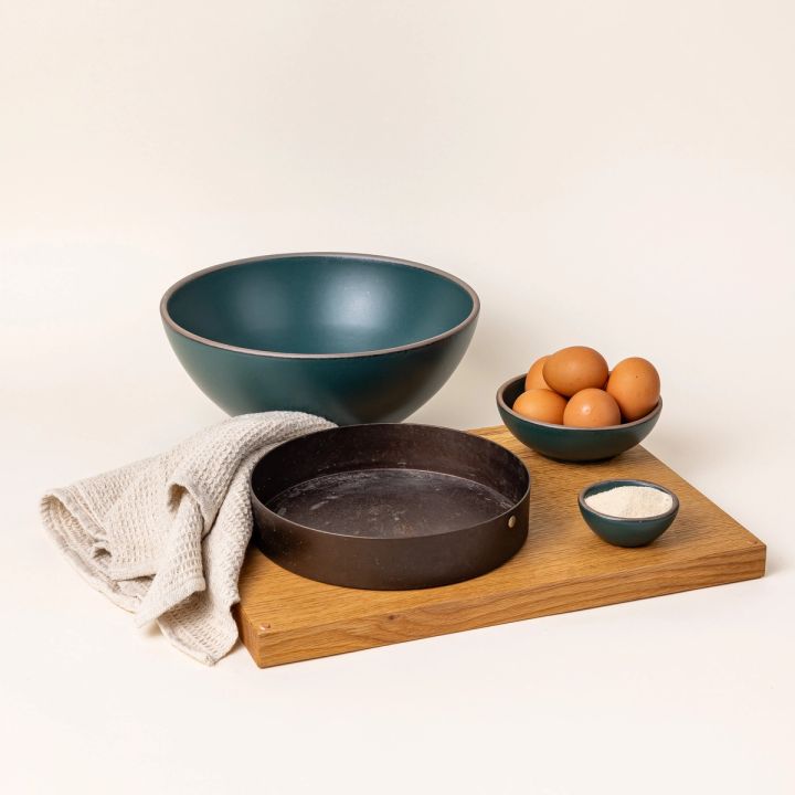 A styled baking set up with a dark teal mixing bowl, a cake pan, a kitchen towel, a bowl full of eggs, and a tiny bowl full of flour. All items are on or near a large light wood cutting board.