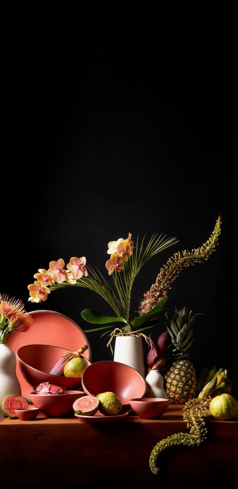 Watermelon pink ceramics with tropical fruits and flowers arranged on a table against a black background in a vibrant, dramatic display.