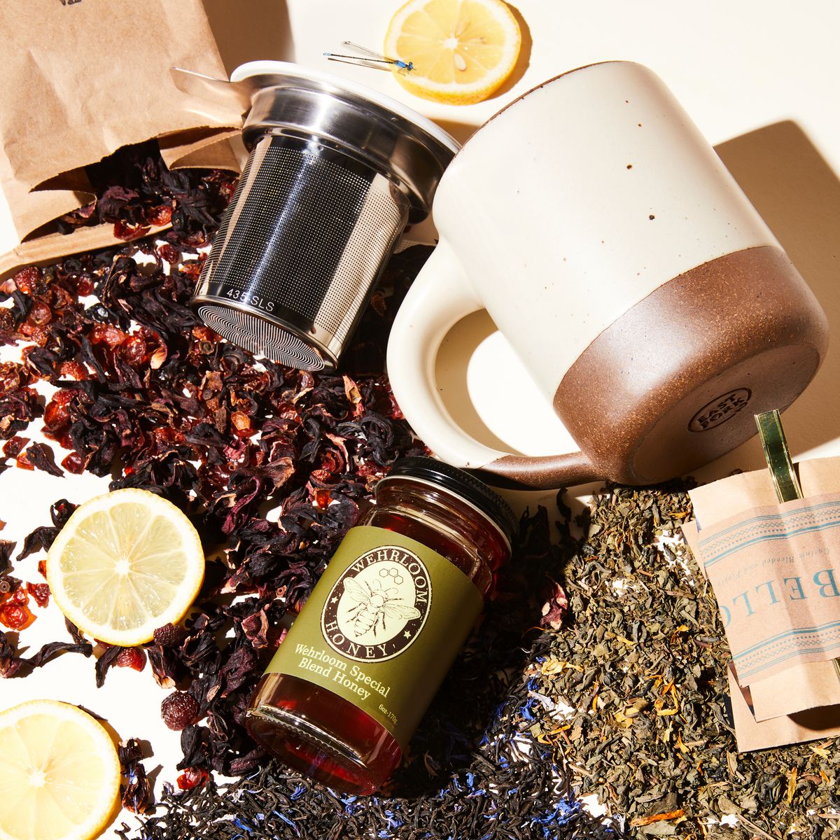 Two open bags of tea with three colorful varieties of loose tea, cut lemons with a jar of honey, big panna cotta mug, and metal tea strainer lying on their sides