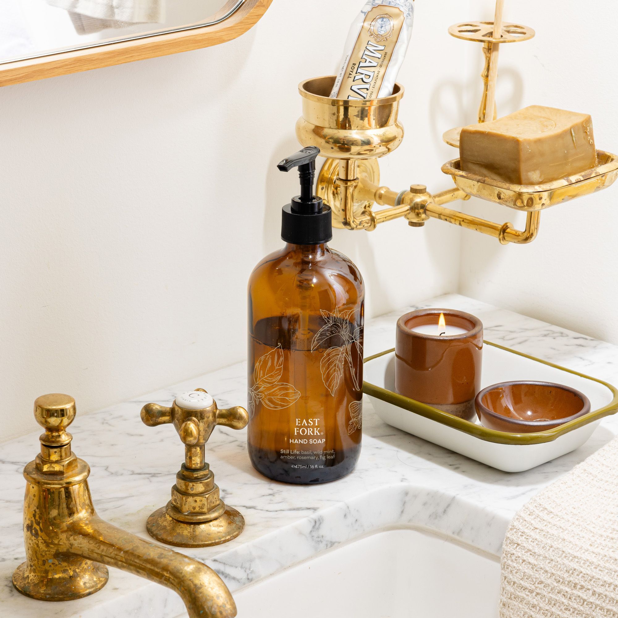 Amber hand-soap bottle on a marble sink beside brass fixtures, a cool terracotta ceramic candle, and a tray with small terracotta dishes in a bright bathroom.