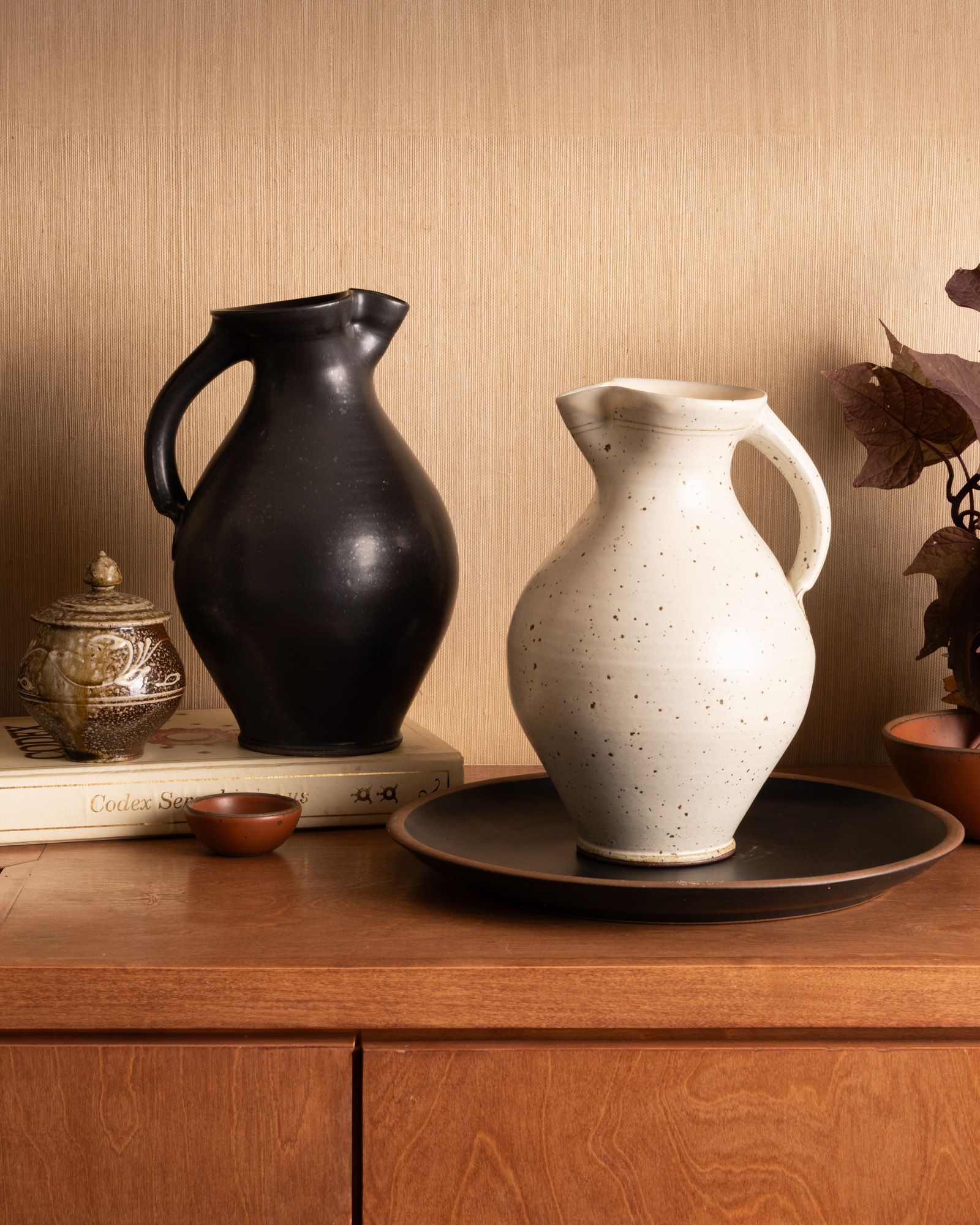 Black and cream ceramic pitchers on a wooden surface, styled with books, bowls, and foliage against a neutral wall.