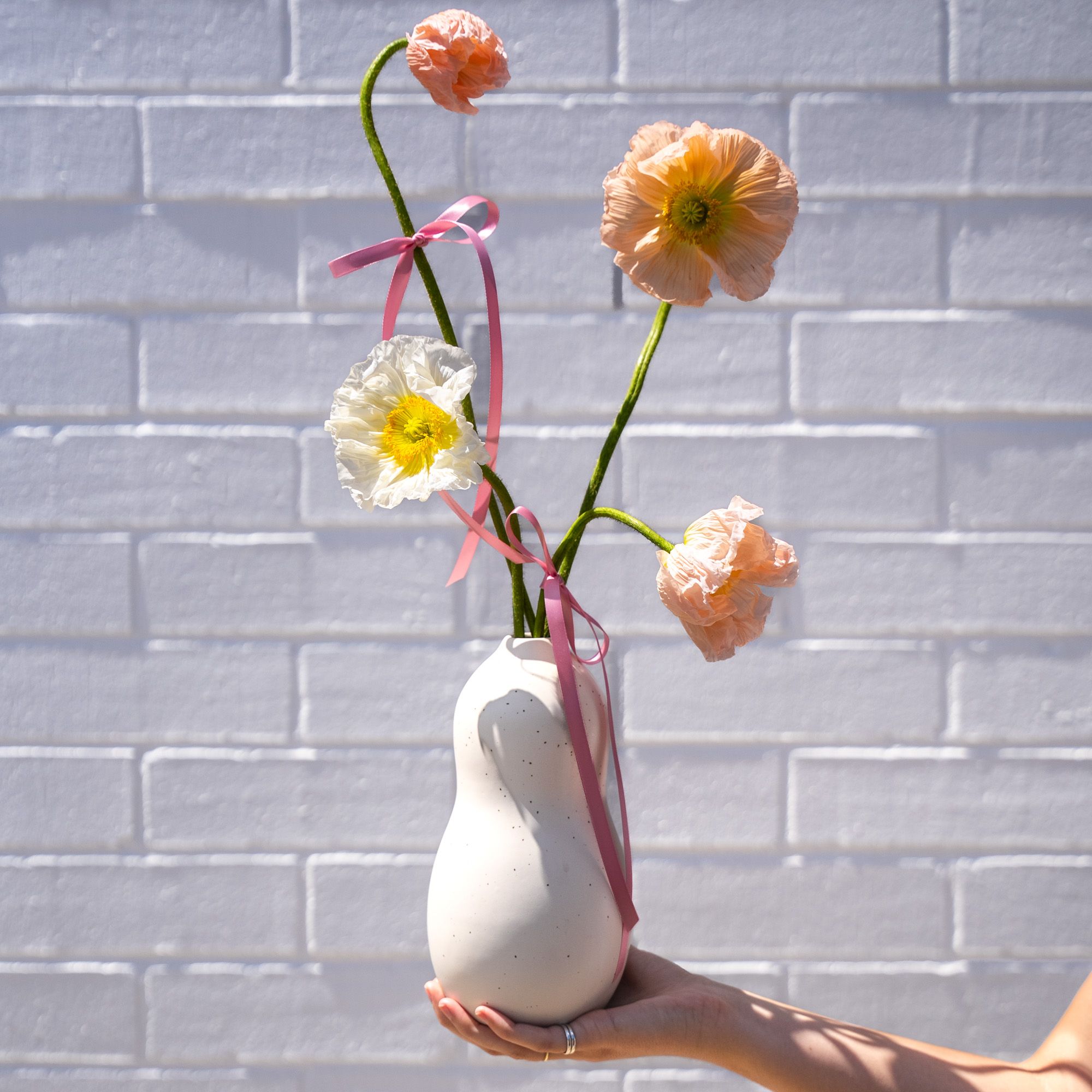 A hand holds out a tall, cream-colored ceramic vase holds pastel poppies in shades of peach, white, and orange, each stem adorned with pink ribbons, set against a white brick wall.
