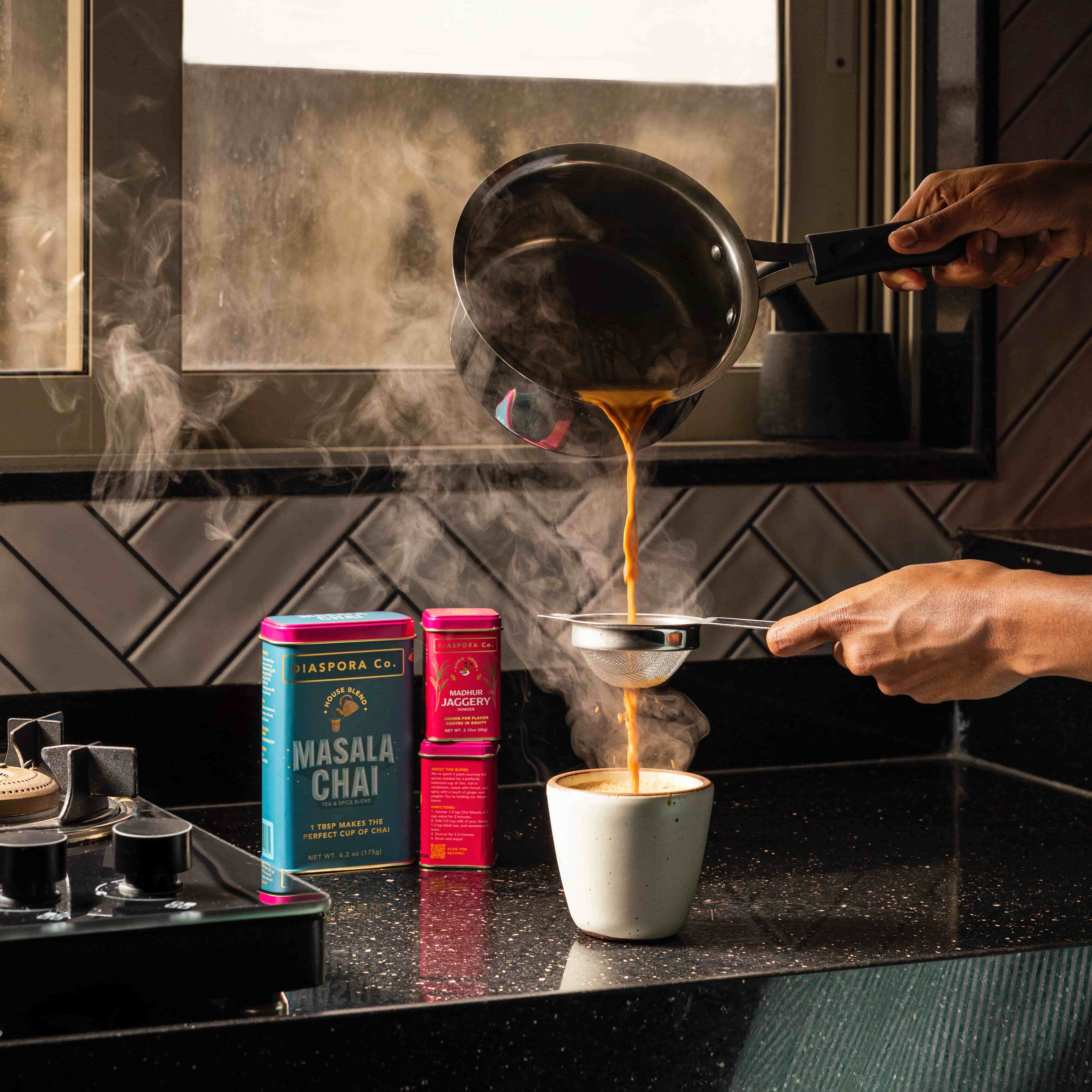 On a kitchen counter, a hand holds a strainer over a small ceramic cup pouring chai, next to two food tins of masala chai and jaggery.