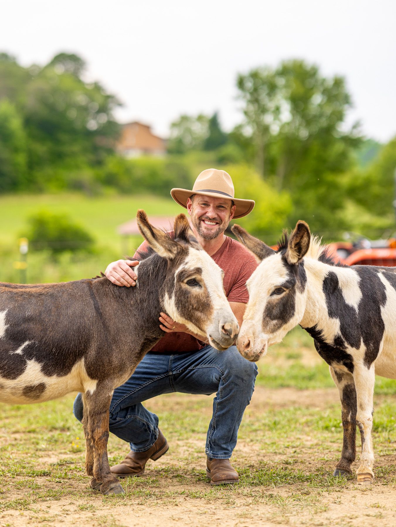 A man is outside smiling at the camera while petting two adorable donkeys.