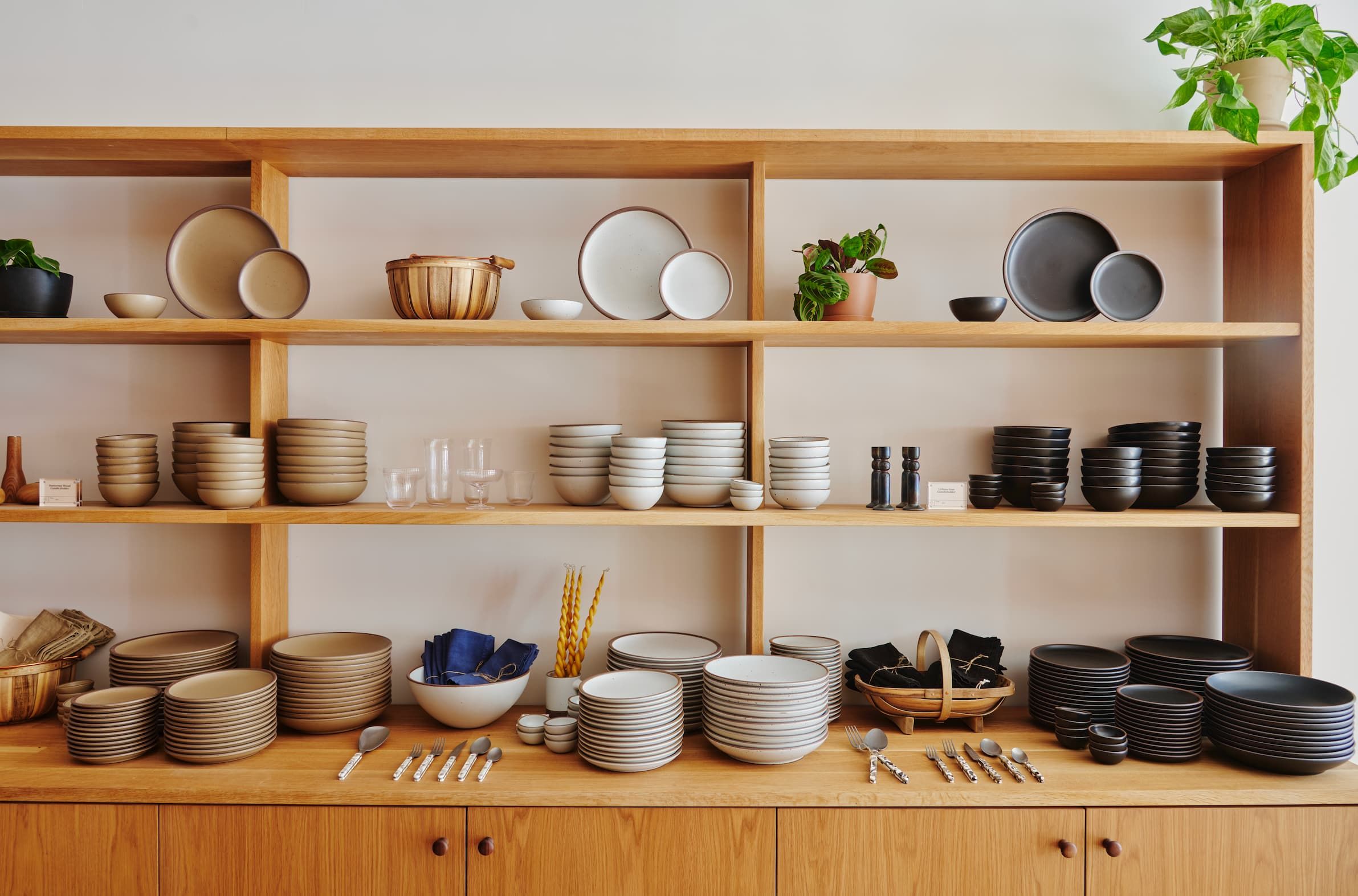 Warm wood shelves highlight neutral tan, white and black ceramic dinnerware with flatware, napkins, candles and more merchandised as well.