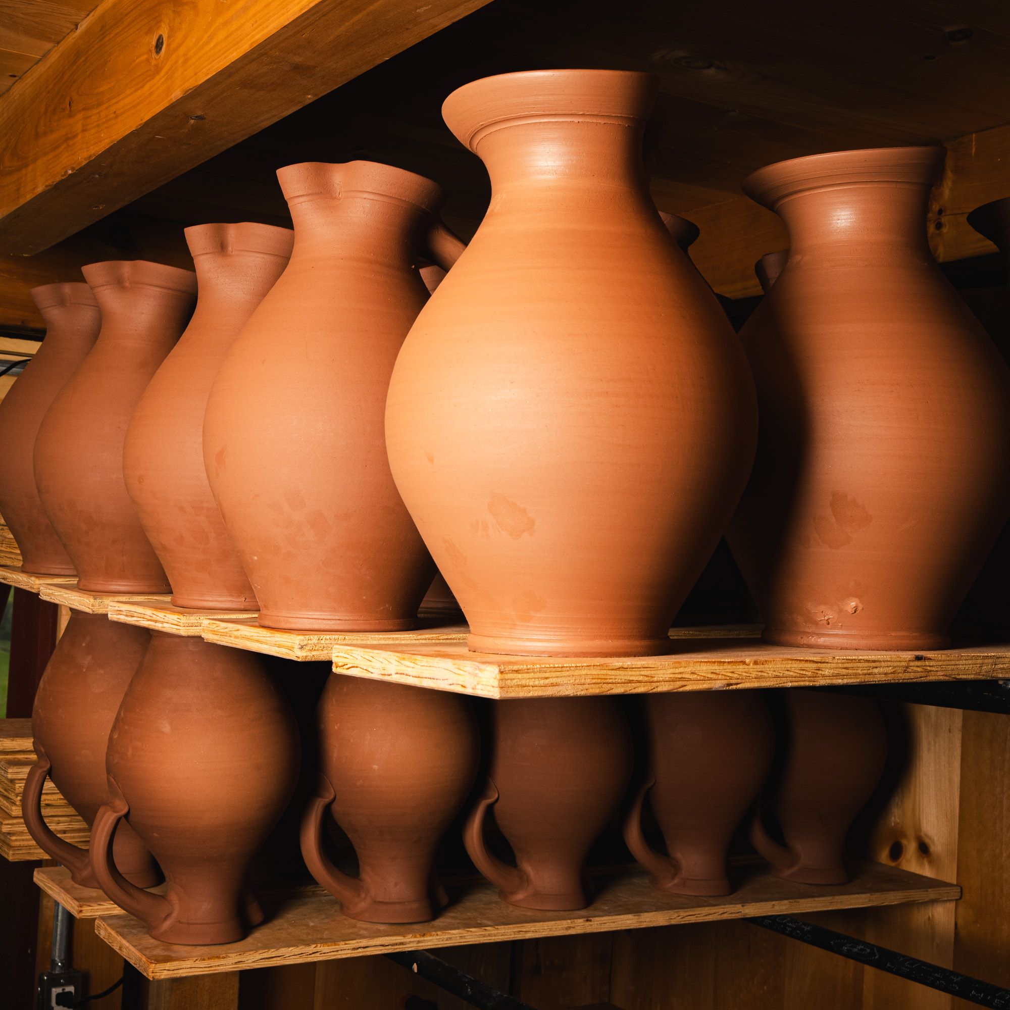 Rows of unglazed formed pitchers drying on wooden shelves in a pottery studio.
