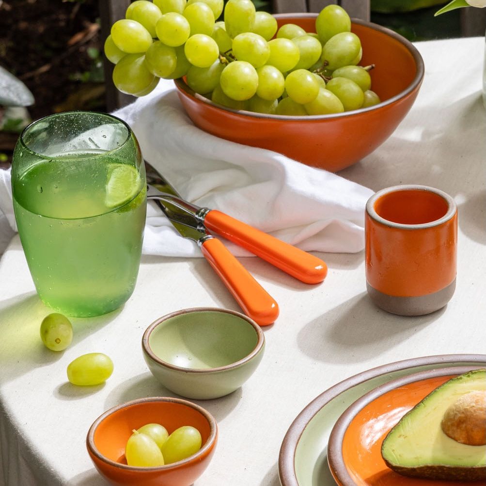 On a table is ceramic plates, bowls, and cups in bold orange and soft sage green colors, a sophisticated green glass filled with seltzer, orange-handled fork and knife, green grapes and a sliced avocado.