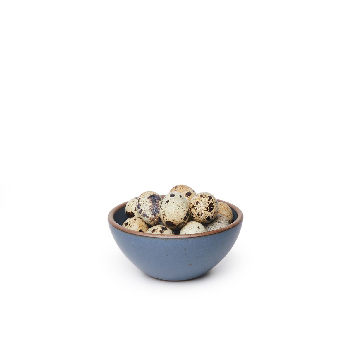 A small dessert sized rounded ceramic bowl in a toned-down navy color featuring iron speckles and an unglazed rim, filled with quail eggs