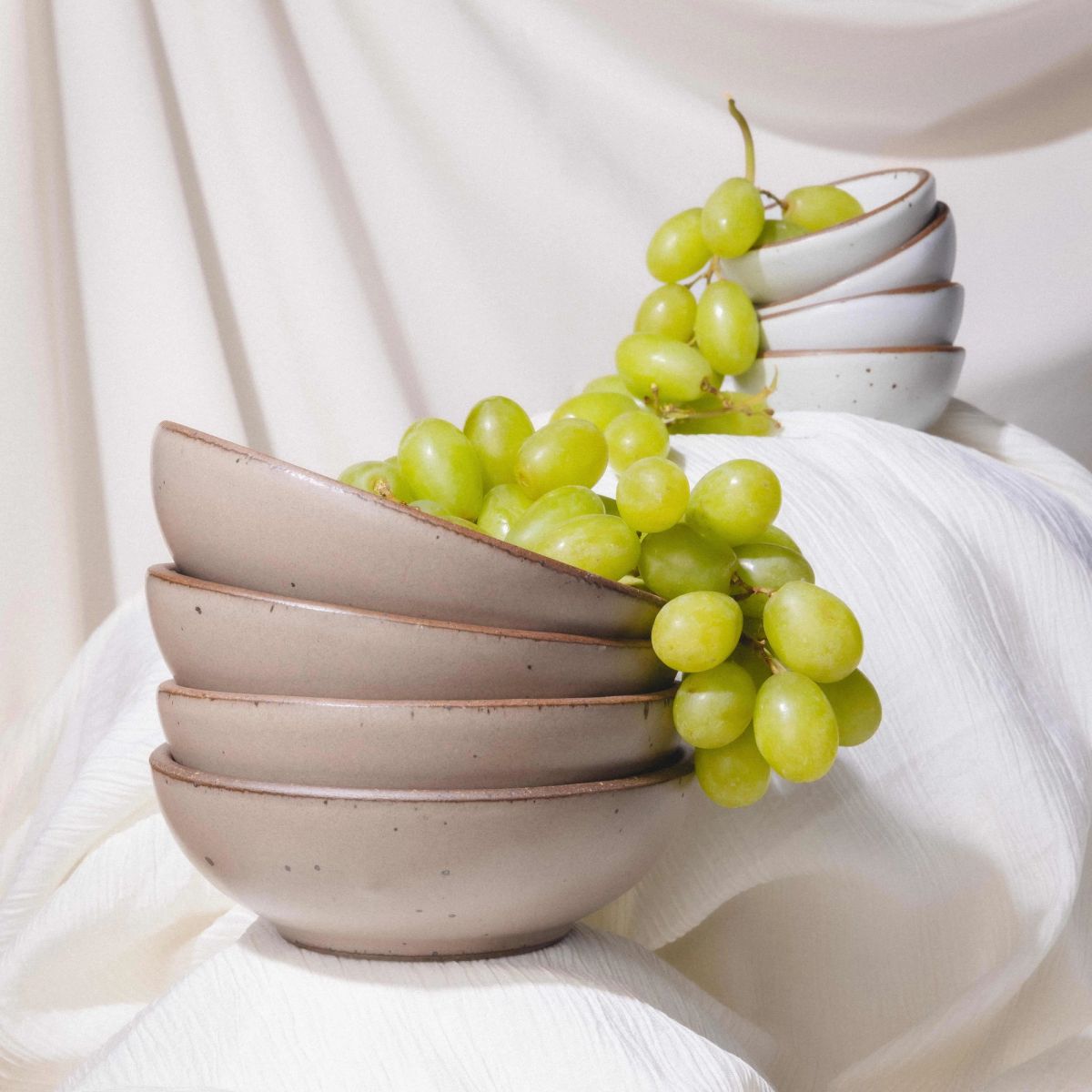 Stacked ceramic bowls with green grapes spilling over, set against soft white draped fabric.