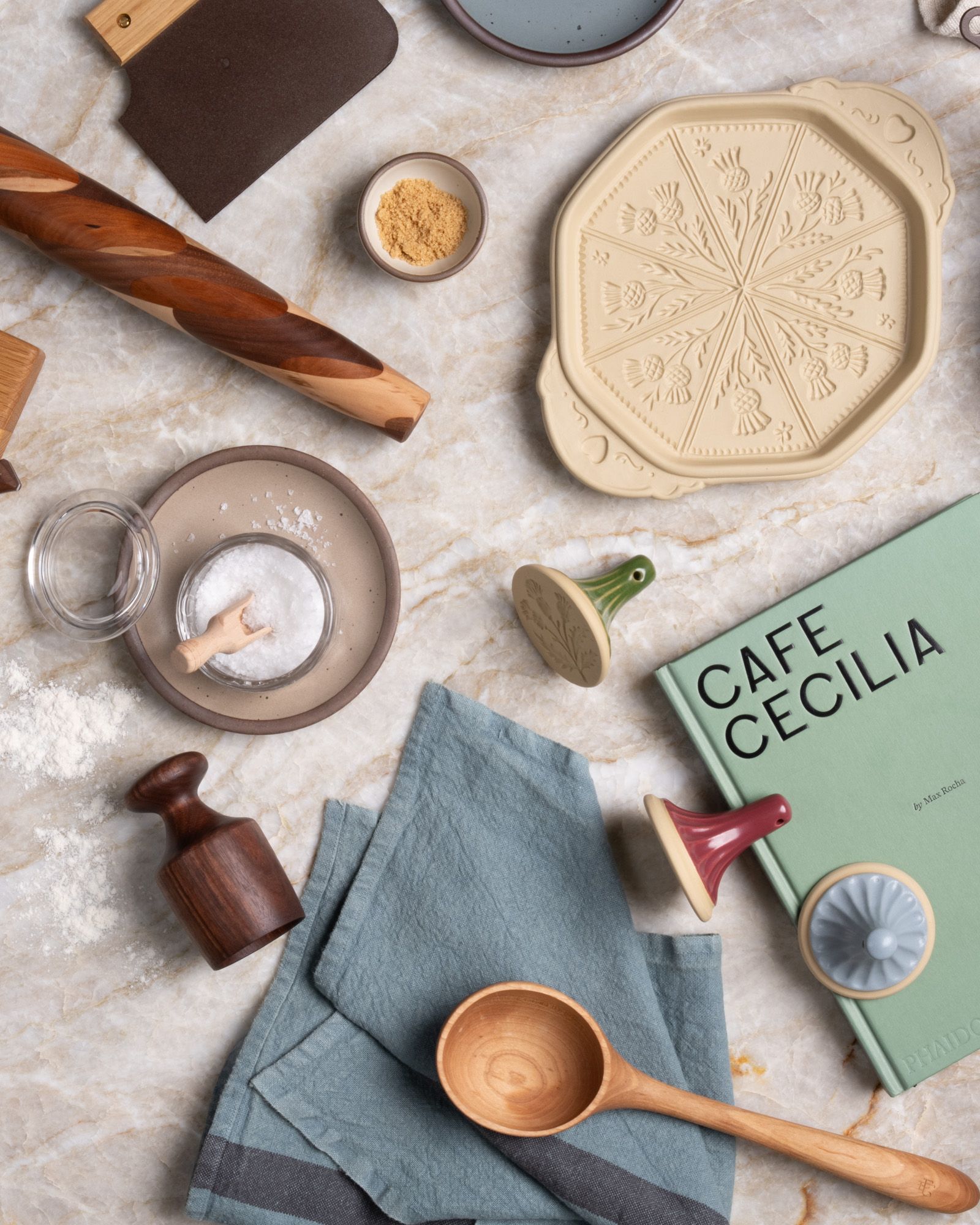 A cozy kitchen scene with wooden utensils, cookie stamps, a biscuit cutter and Café Cecilia cookbook arranged on a marble countertop.