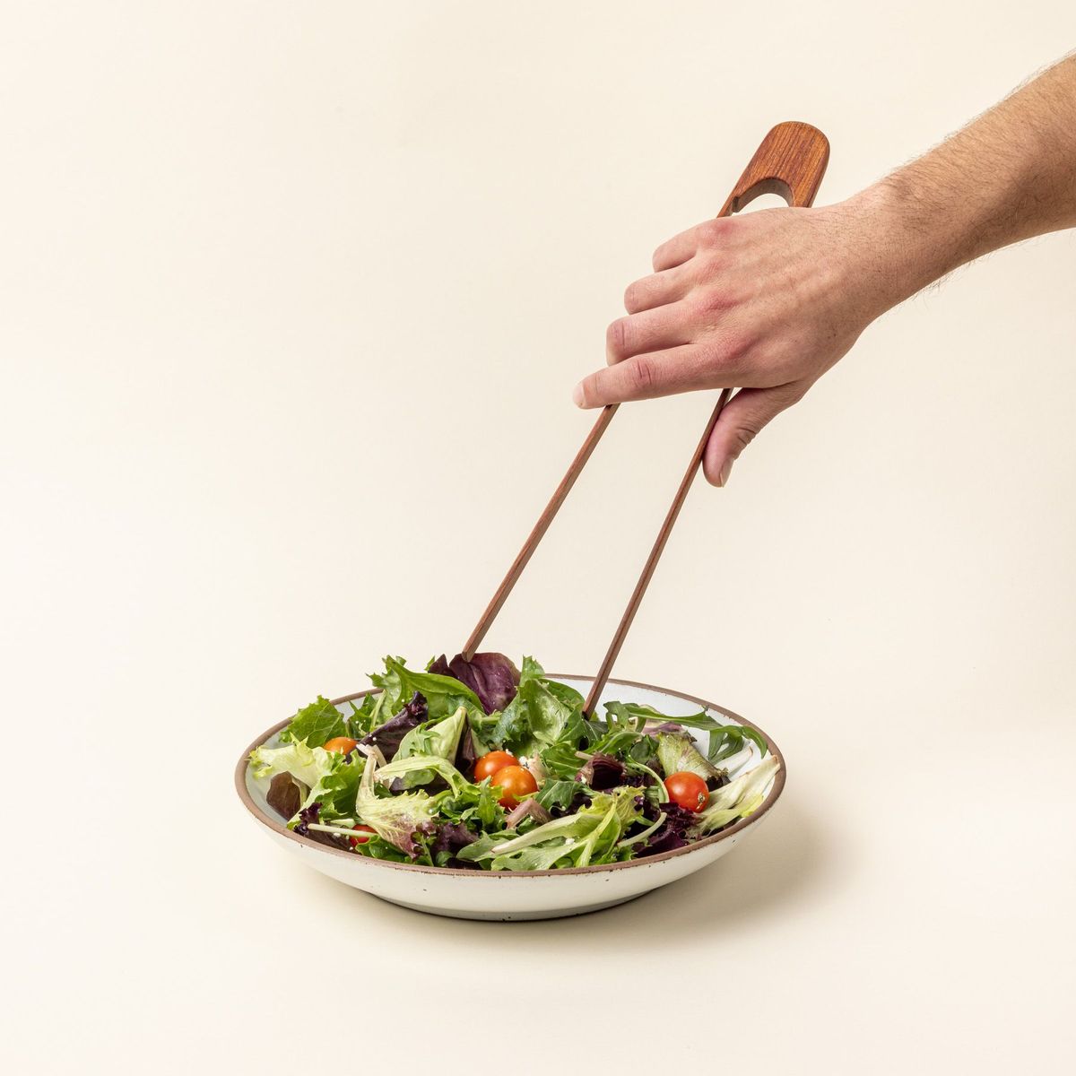 A hand holds simple and minimal wood rectangular tongs reaching to place a small side salad.