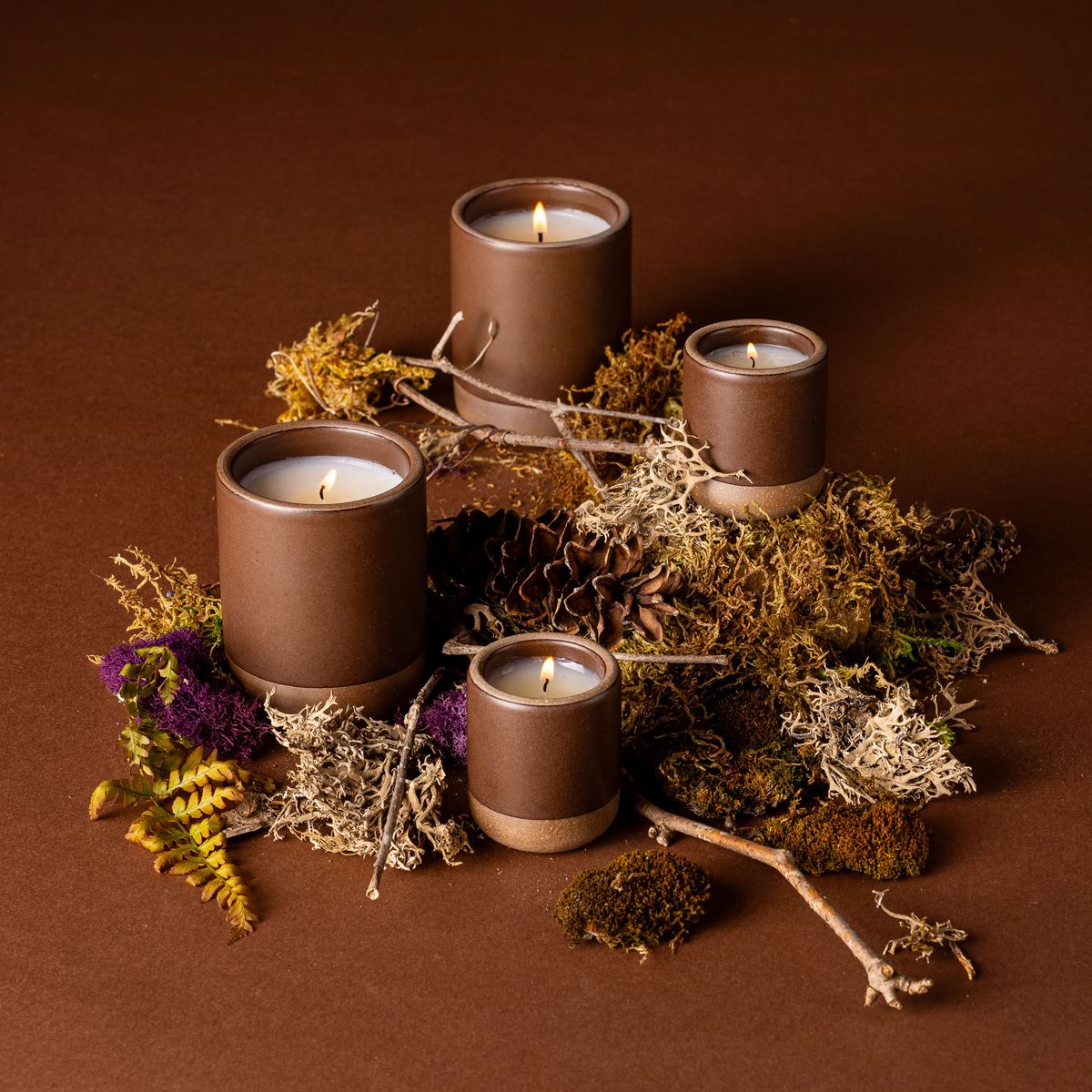 Four lit candles in ceramic vessels in a dark brown color with a match lighting one. They are artfully arranged and surrounded by dried grass, branches, and fern leaves.