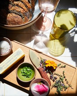 A board filled with herbs, a stick of butter, a knife, and a bowl of sliced bread along with colorful glasses.