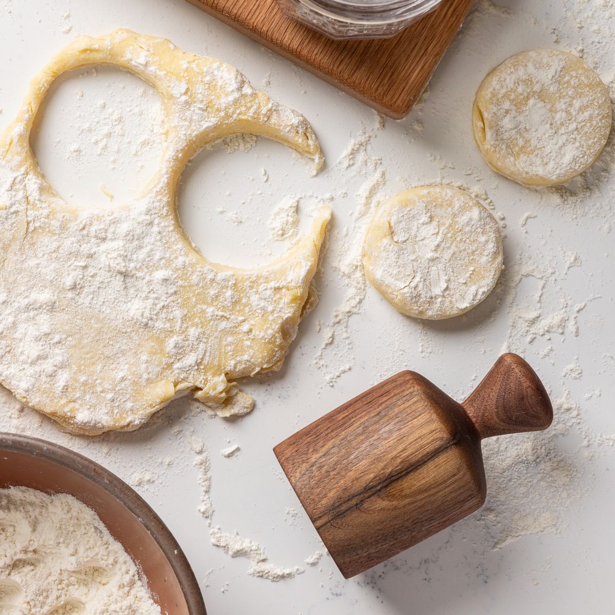 On a white countertop, there is a wooden cylindrical black walnut biscuit cutter with a turned knob, cut out biscuits, flat dough and sprinkled flour, and a bowl filled with flour.