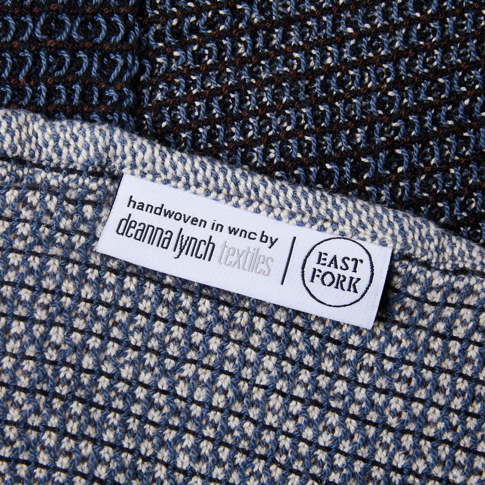 Close up of a woven blue, black, and white blanket with a white label that reads "Handwoven in WNC by Deanna Lynch Textiles" next to the East Fork logo