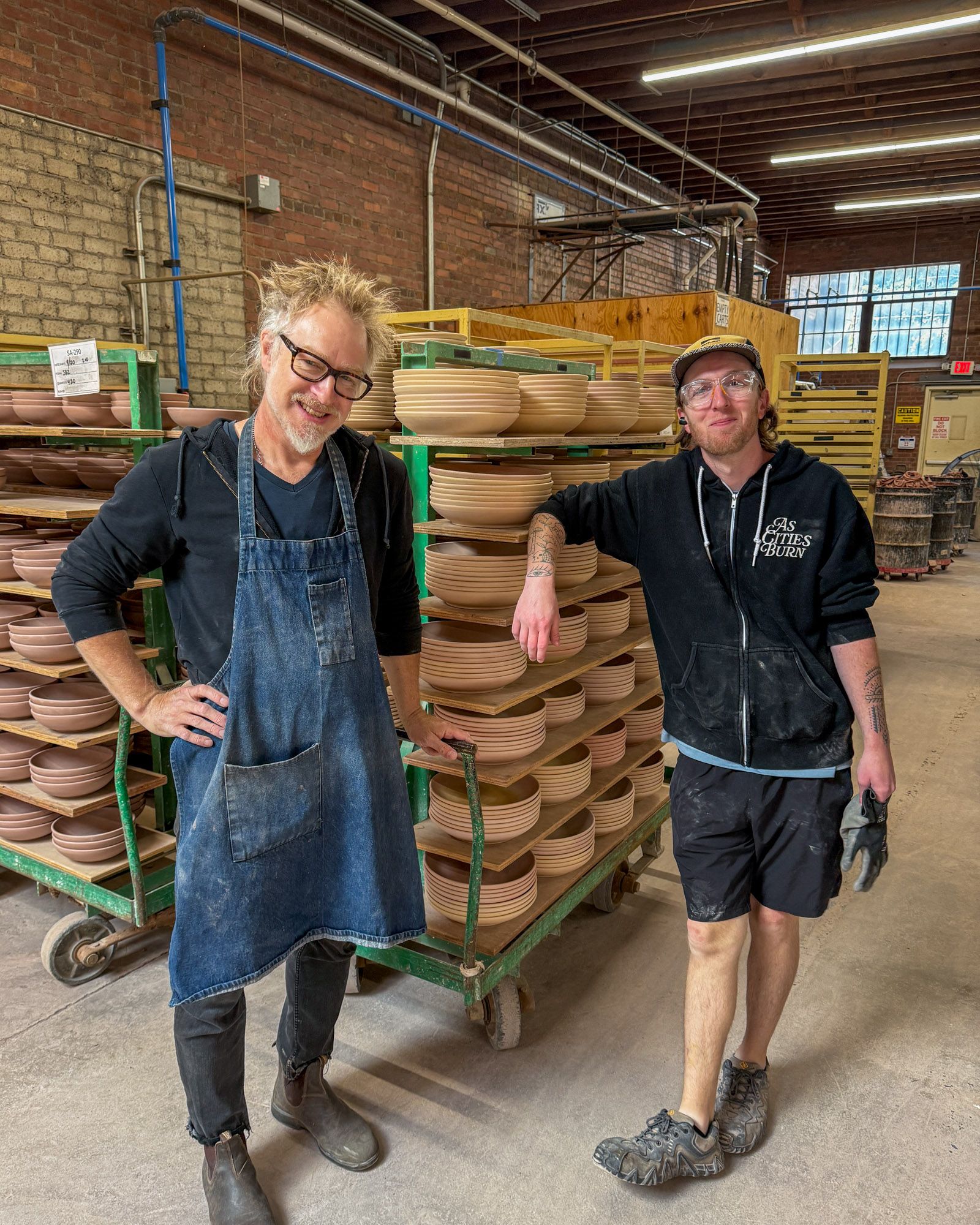 Two East Fork employees are back in the studio and smiling in front of carts of unfinished pottery