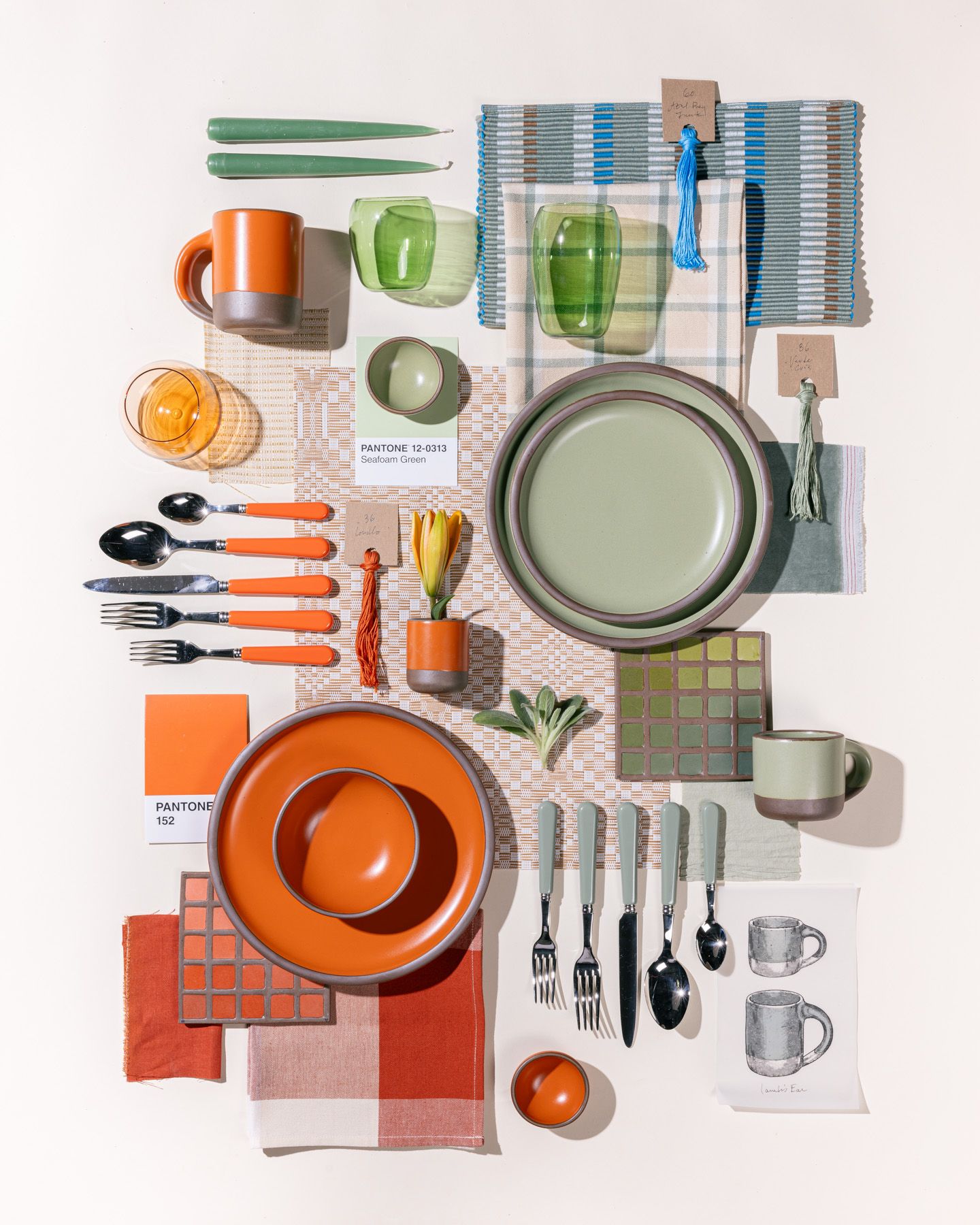 An overhead view of an artistic flat lay of ceramic plates in sage green and bold orange colors, surrounded by pantone cards, textile samples, flatware, glassware all in matching colors.