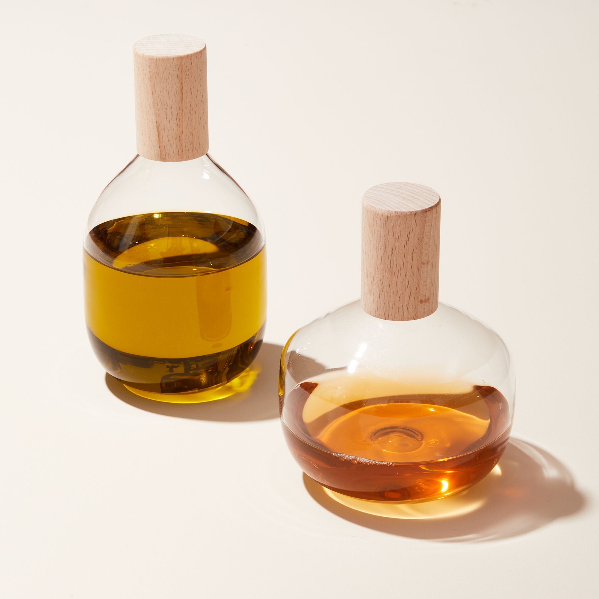 2 Glass olive oil cruets with cylindrical wood tops. The cruet on the left is tall and wide, the cruet on the right is short and wider. Each are filled with a type of cooking oil.