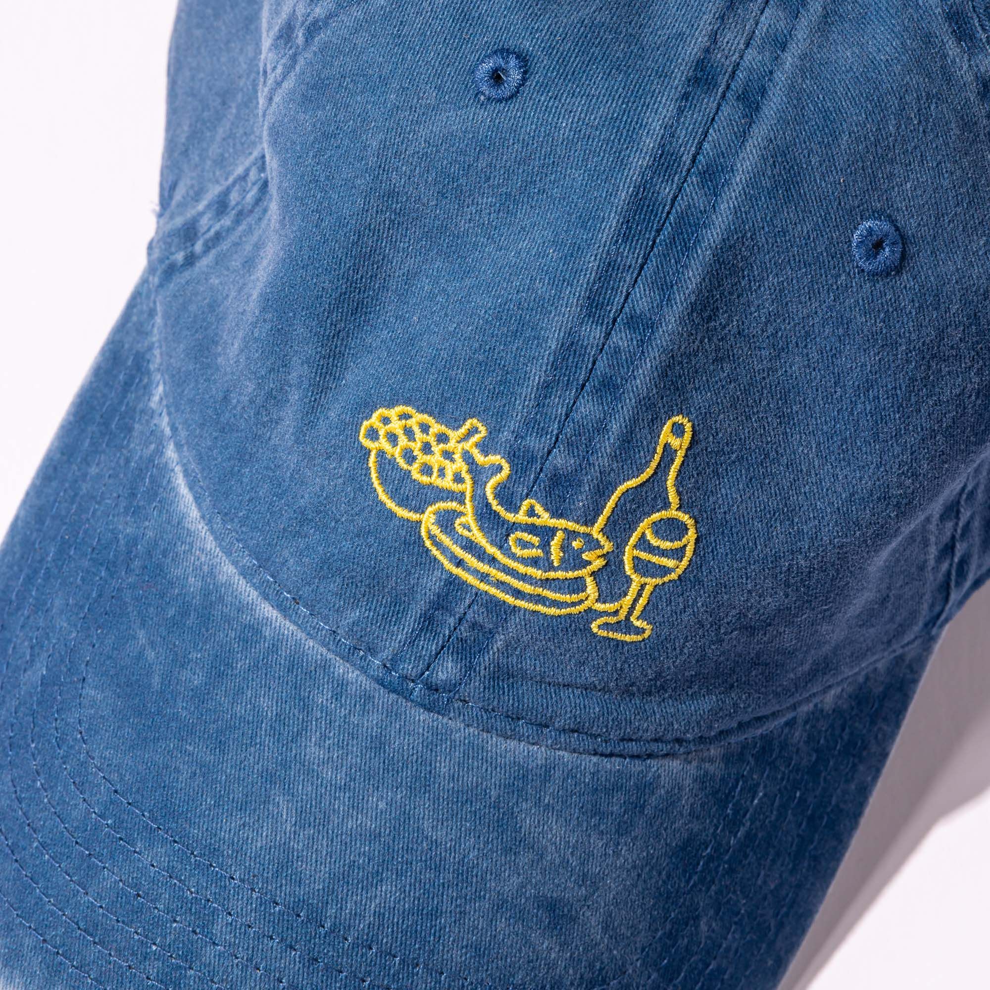 A close up of the classic blue baseball hat with yellow embroidery on the front depicting a feast of fish, fruit and wine.