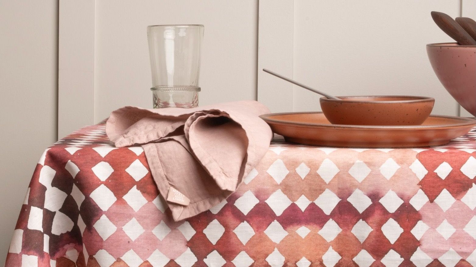 Table set with pink ceramic dishware on a watercolor diamond patterned tablecloth, with a wooden chair and linen napkin nearby.