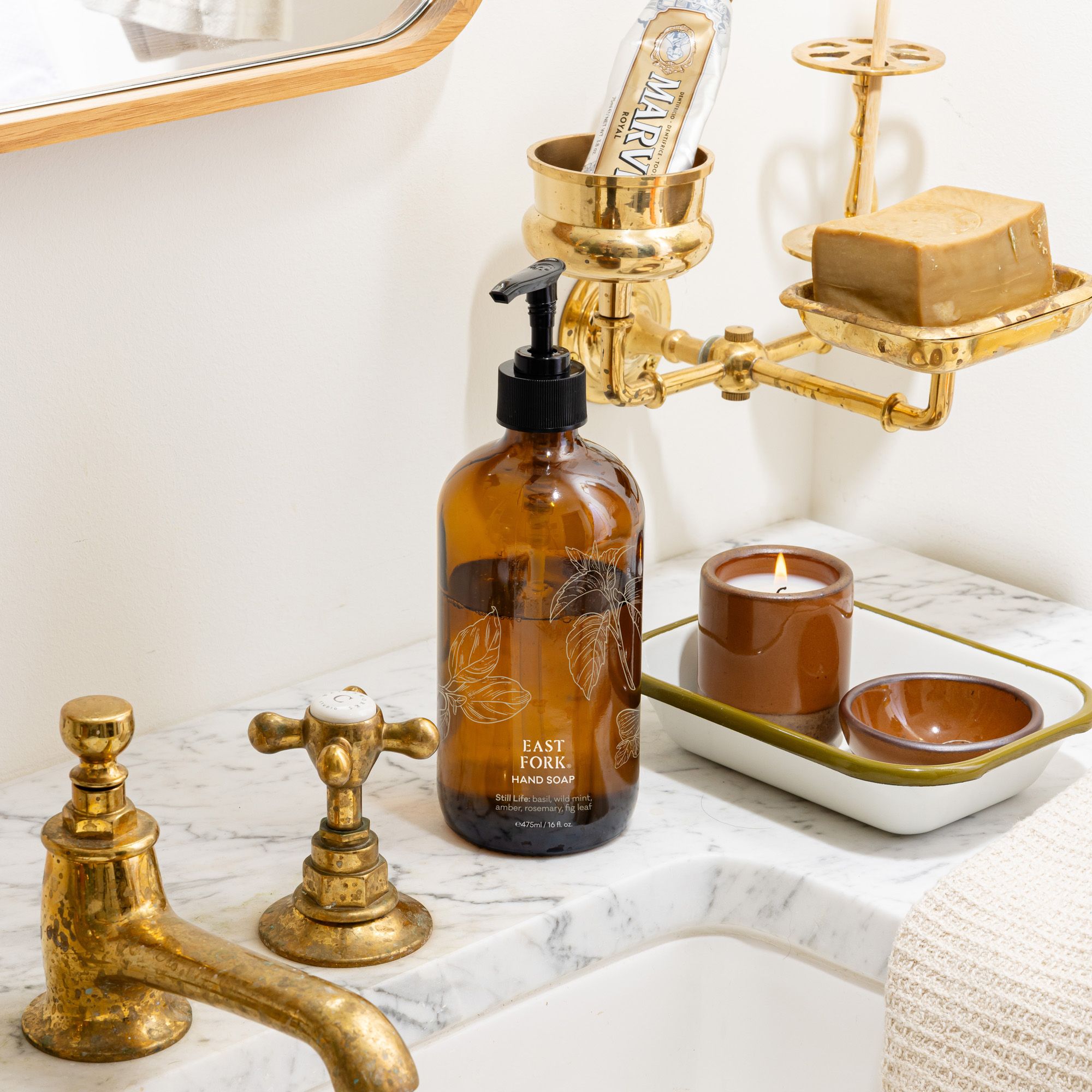Amber hand-soap bottle on a marble sink beside brass fixtures, a cool terracotta ceramic candle, and a tray with small terracotta dishes in a bright bathroom.