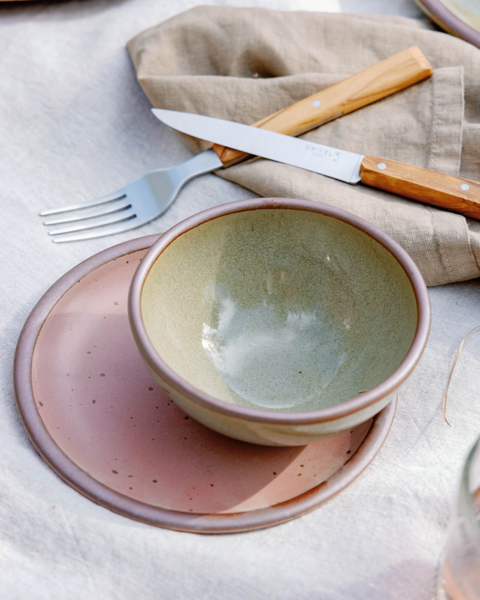 Reflective dappled grey-green bowl and dusty mauve pink side plate with wood handled fork and knife outside at a picnic.