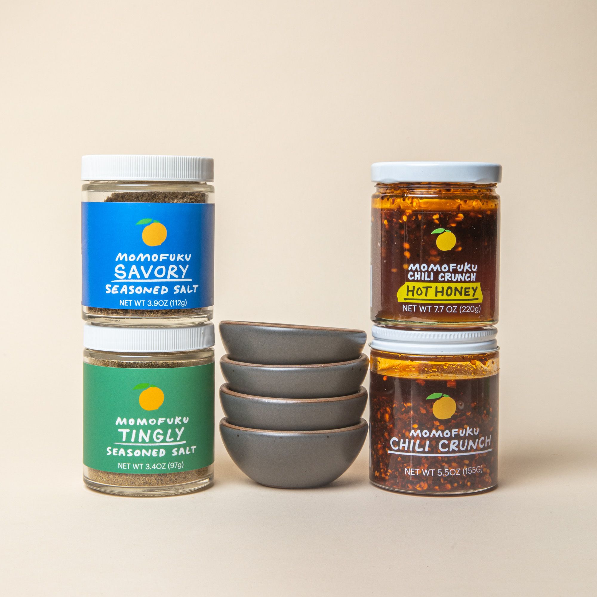 4 little stacked ceramic bowls in a soft cool grey color surrounded by 4 jars of Momofuku seasoned salts and chili crunch.