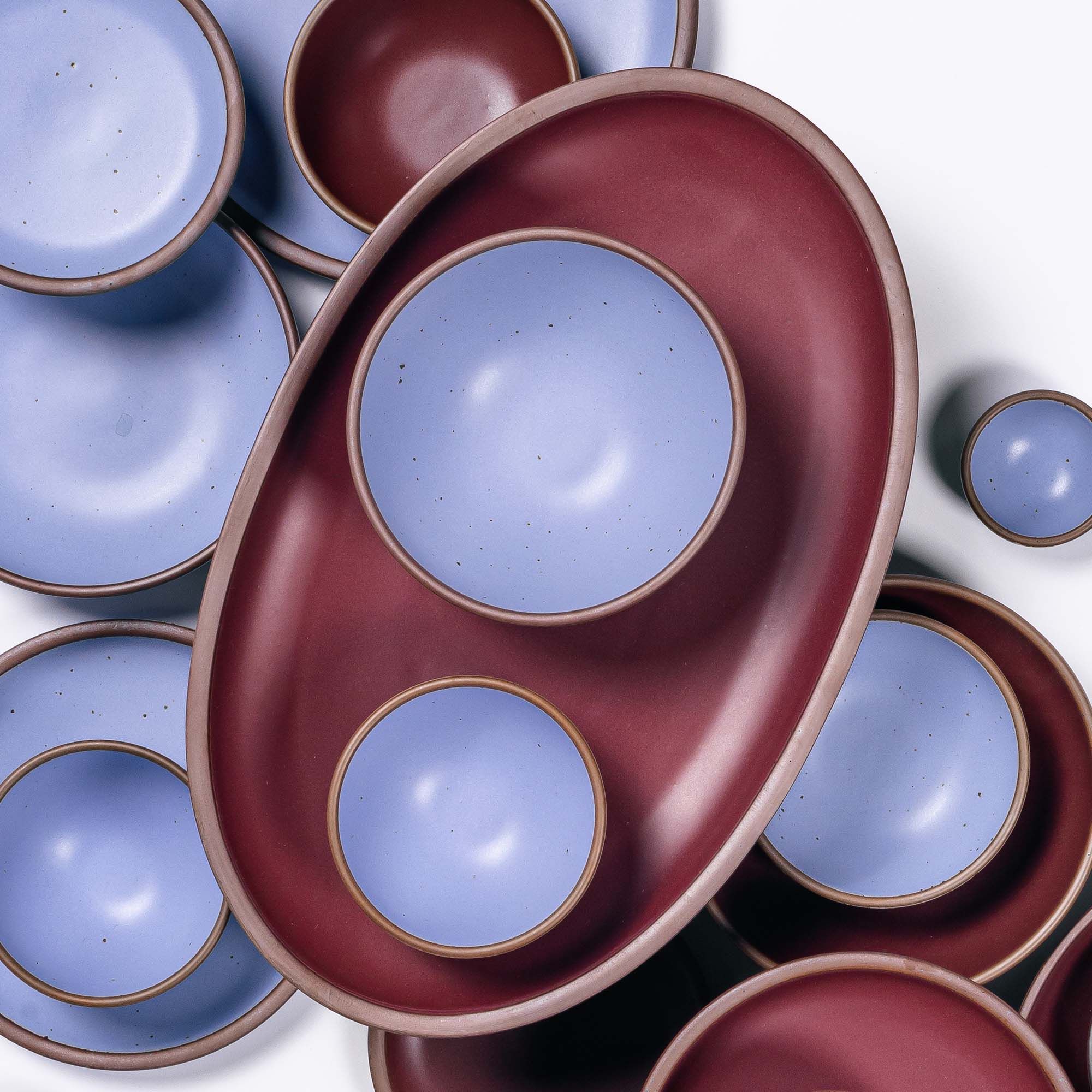 A large oval ceramic platter in a plum color is surrounded with smaller ceramic plates and bowls in periwinkle