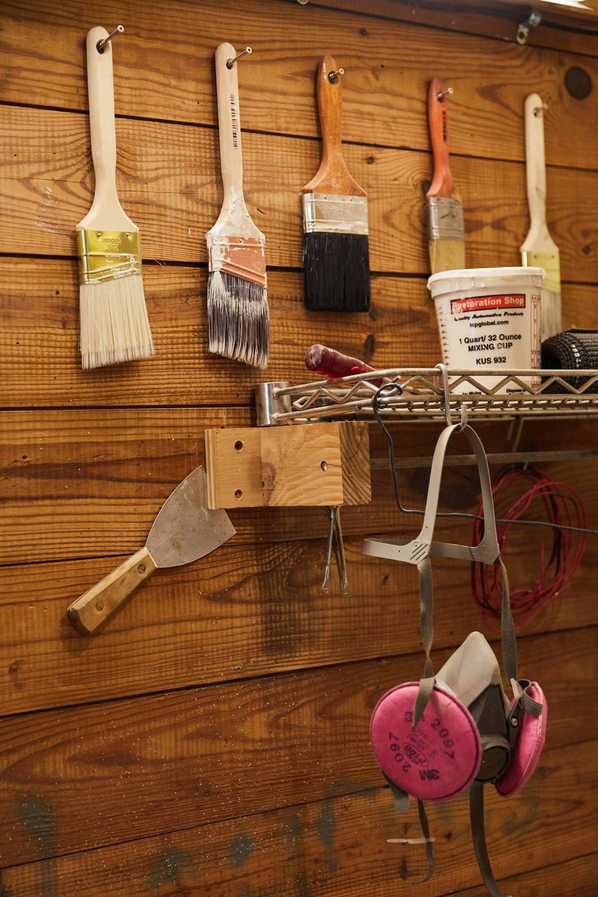 Some tools hang on the wall of woodworking workshop, including wide paint brushes, a mask, and more