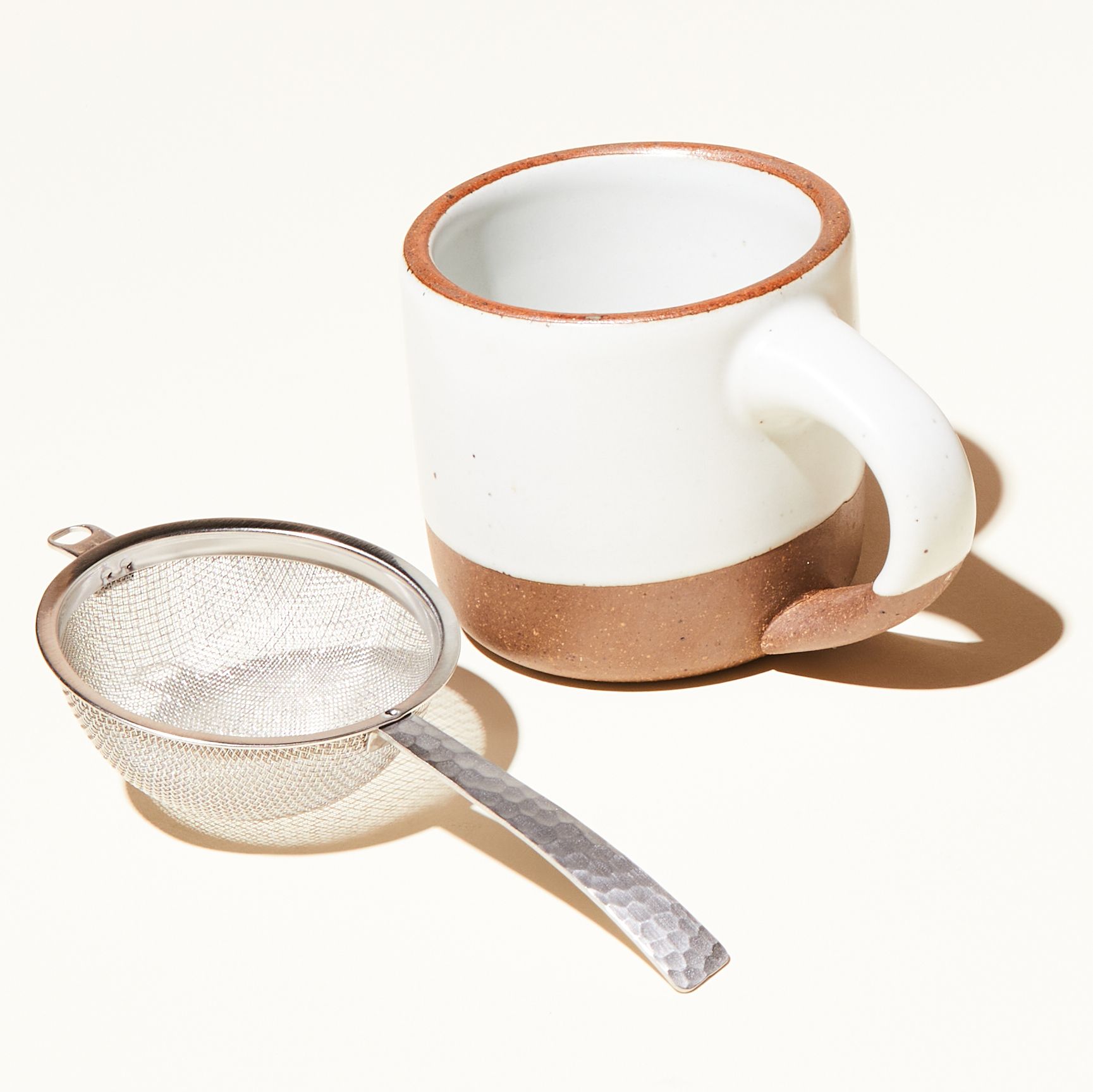 Stainless steel half dome strainer with steel handle laying next to The Small Mug in Eggshell