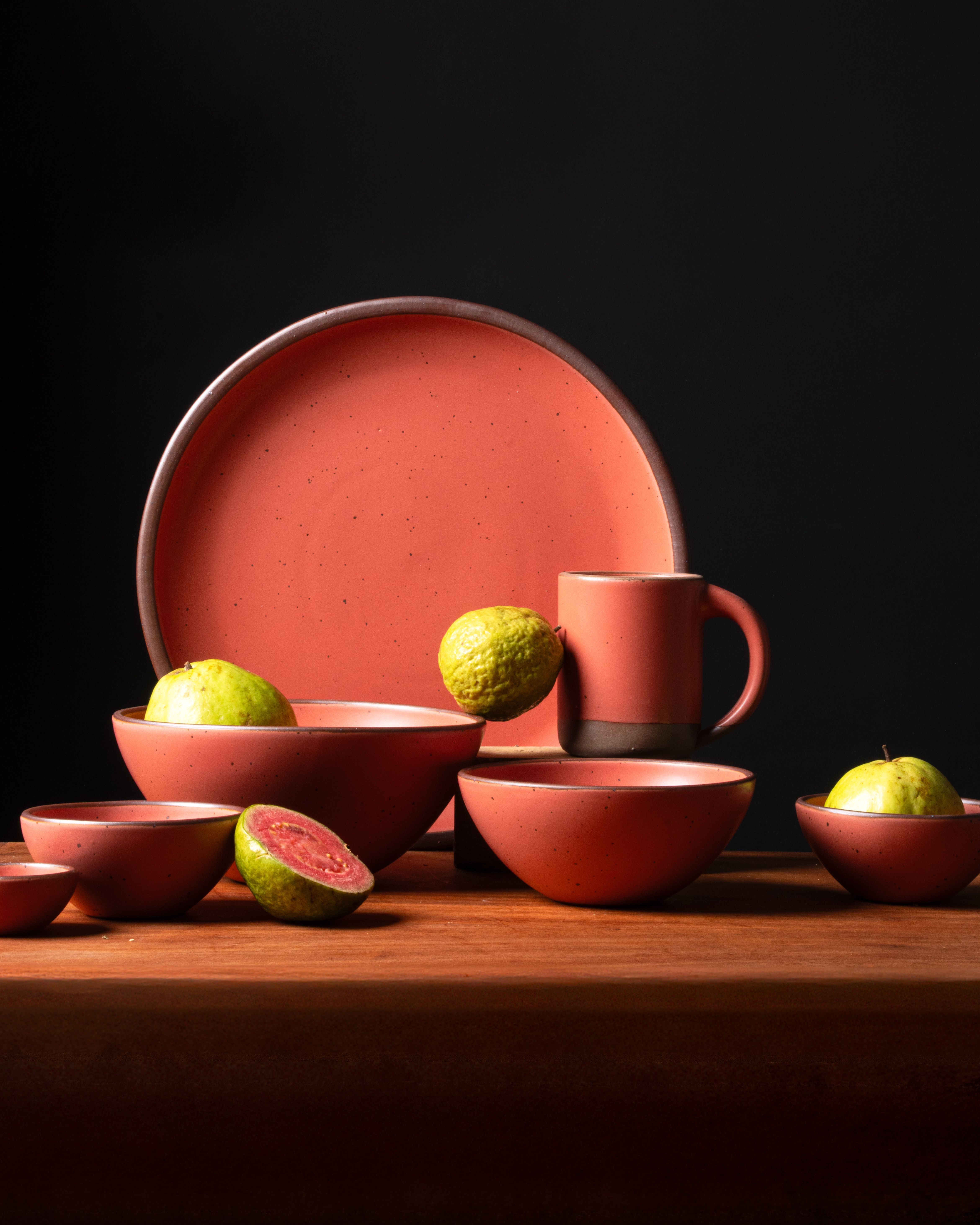 Guava-colored ceramic bowls and plate styled with fresh guavas on a wood surface against a black background.