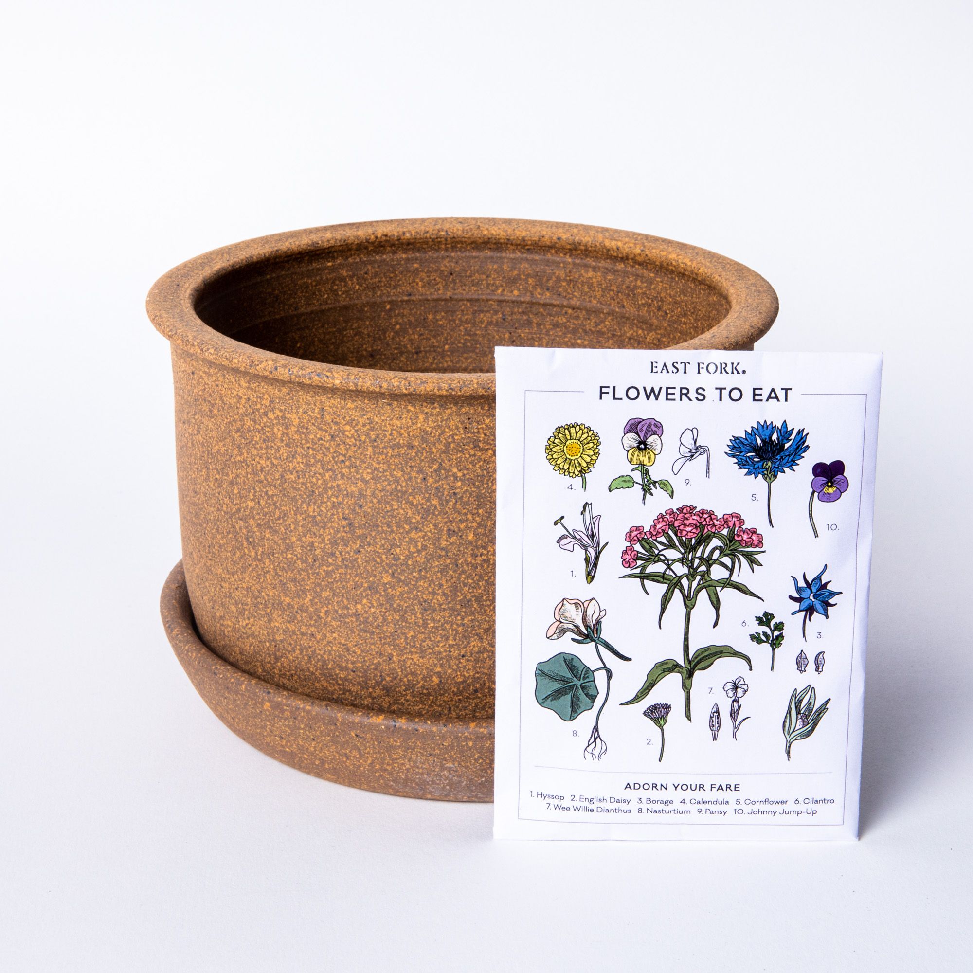 A warm earthy brown ceramic East Fork Workshop planter with a white rectangular bag of flower seeds with floral illustrations on the packaging