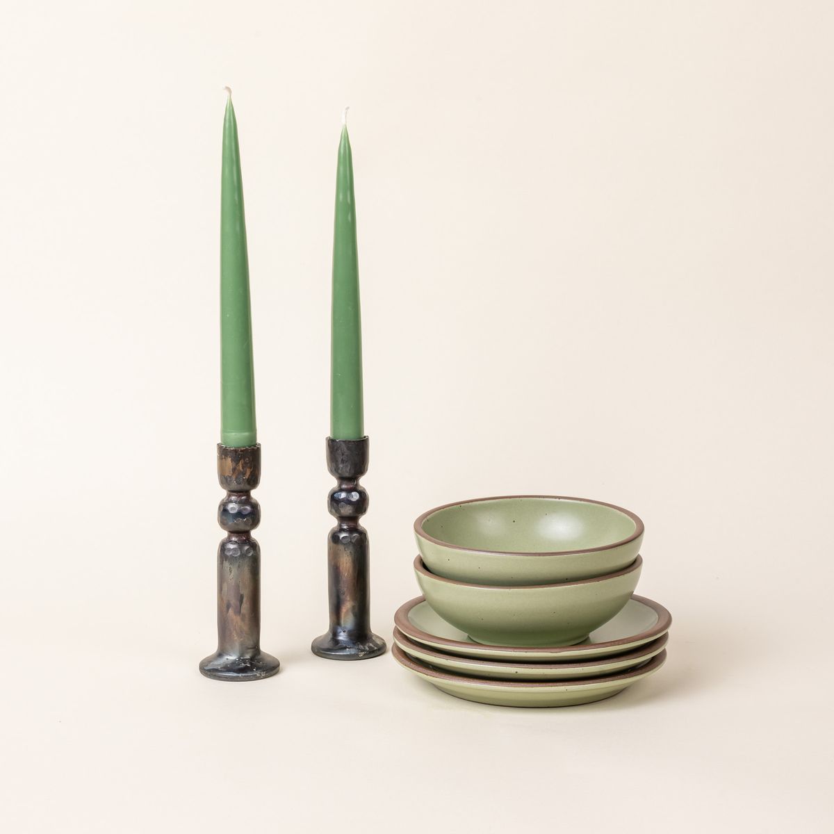 A pair of green taper candles sitting in iron candlestick holders, next to a small stack of sage green bowls and plates.