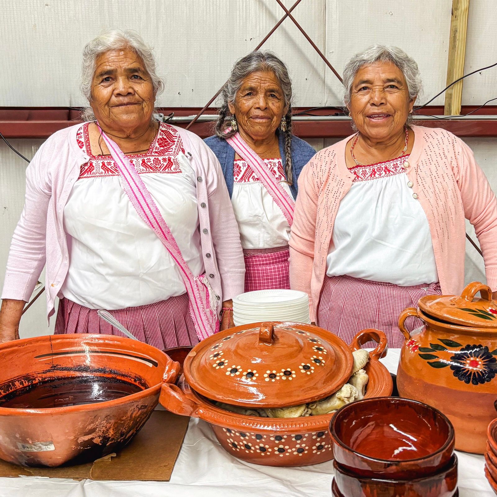 Three people wearing matching white tops with pink embroidery and cardigans standing close together behind a table of large handmade pottery pieces.