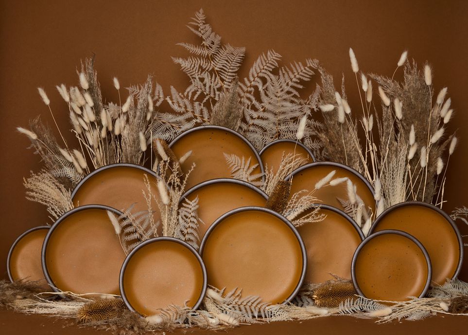 Various sizes of plates in a dark gold color with unglazed rims, surrounded by dried grasses with natural hues