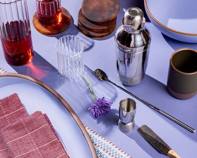 A collection of gifts including a cocktail shaker set, glassware, a mug, and a large oval platter against a periwinkle background.